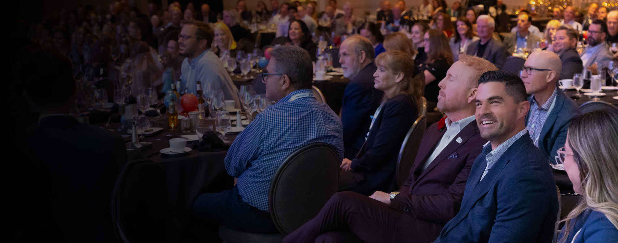 Attendees at a formal conference seated at round tables, listening to a speaker, with festive balloons and drink bottles on the tables.