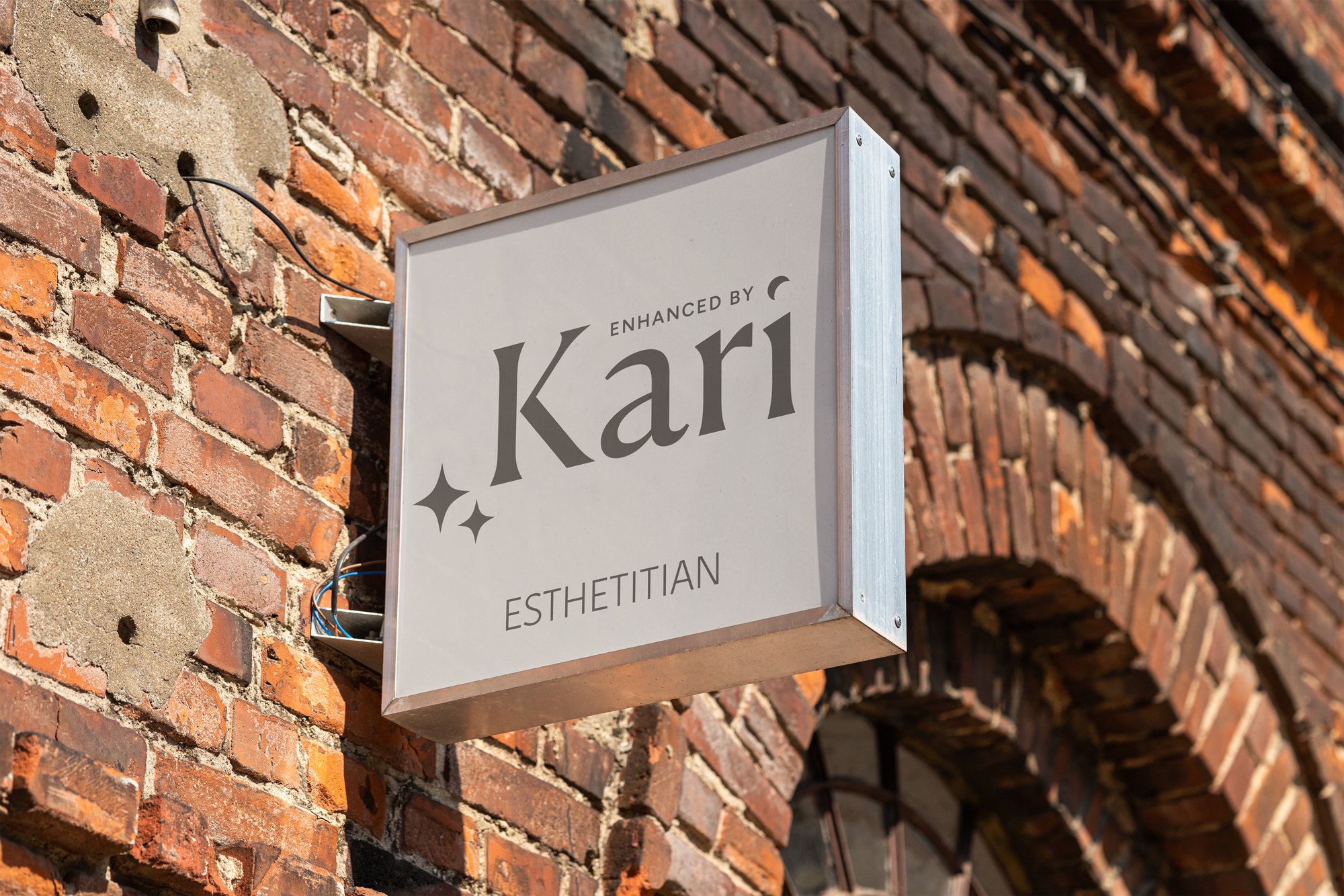 A white sign with gray text and decorative stars reading 'Enhanced by Karl Esthetician' is mounted on a red brick wall.