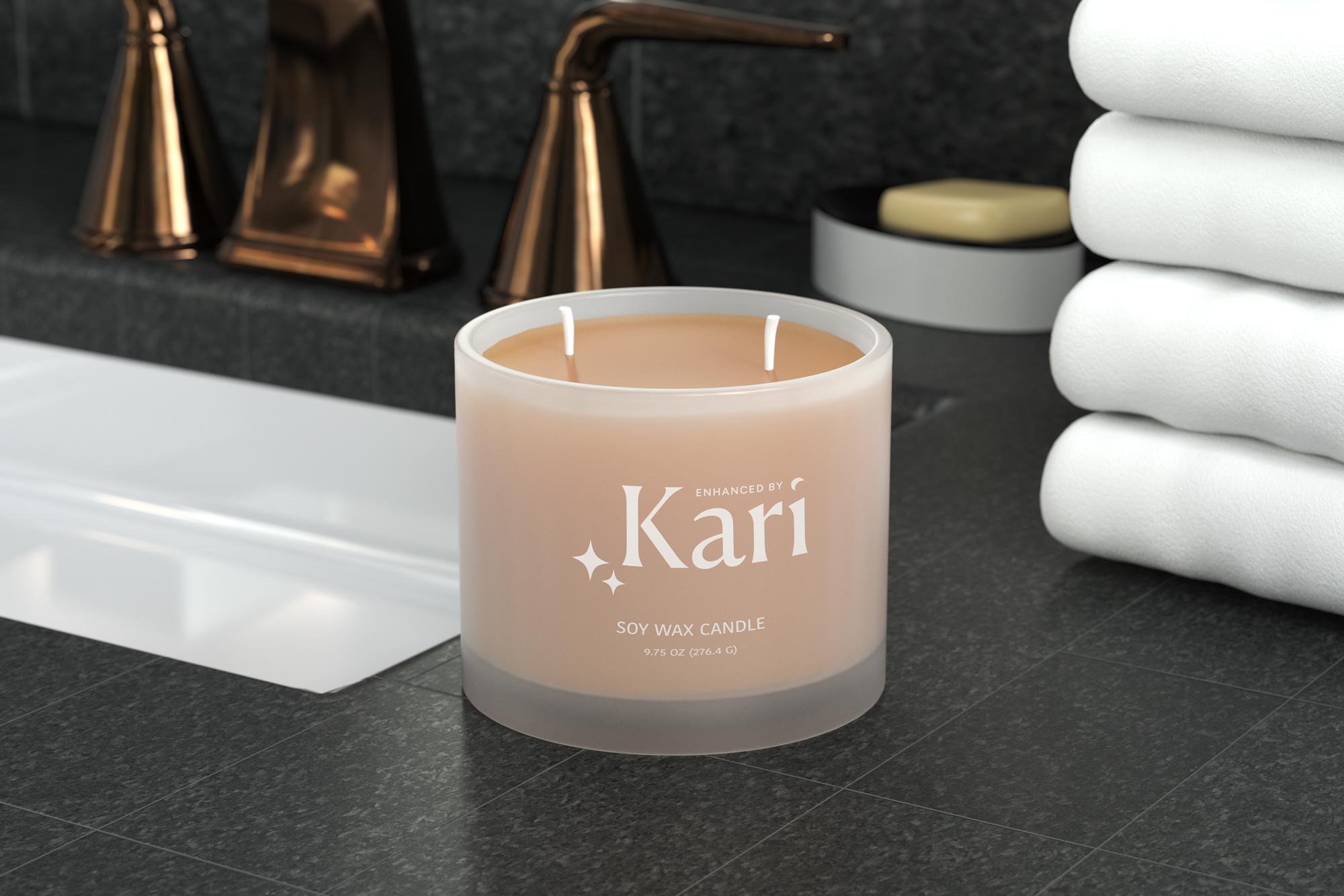 Close-up of a pink soy wax candle labeled 'enhanced by Kari' and 'soy wax candle,' with stacked white towels and a black dish with a beige soap, on a black countertop in a bathroom.