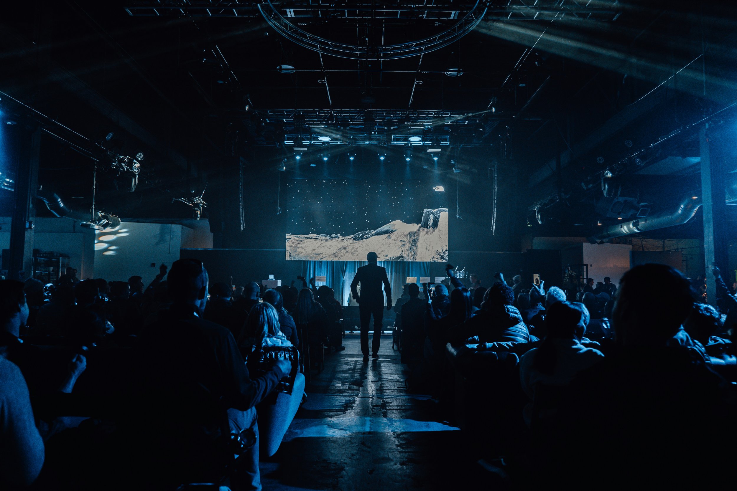 Audience watching a presentation on a large screen in a dark theater, with stage lights illuminating the scene and people seated facing the stage.