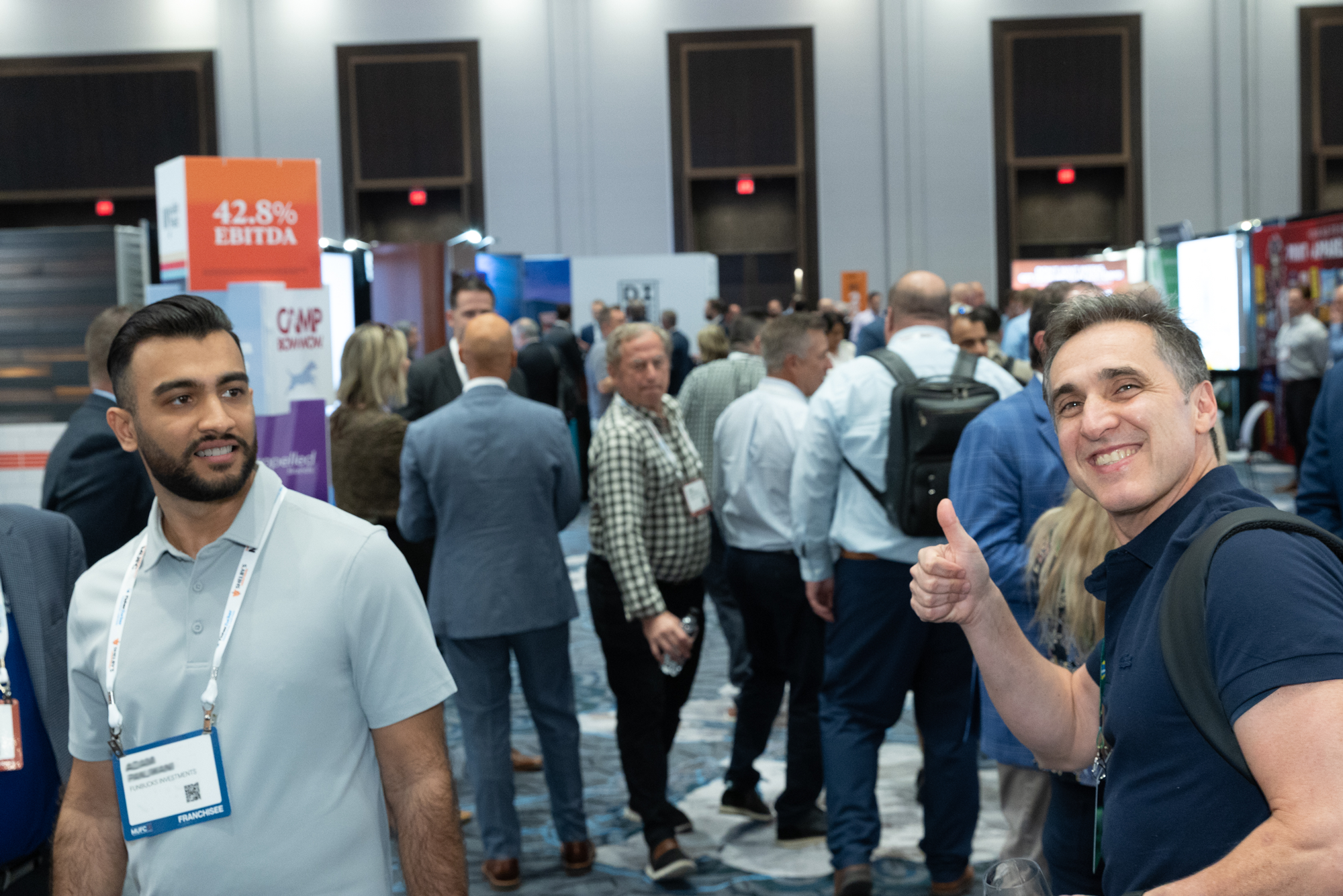 Crowd of people attending a conference or trade show with booths and signage, including a man with a name badge and a man giving a thumbs-up.