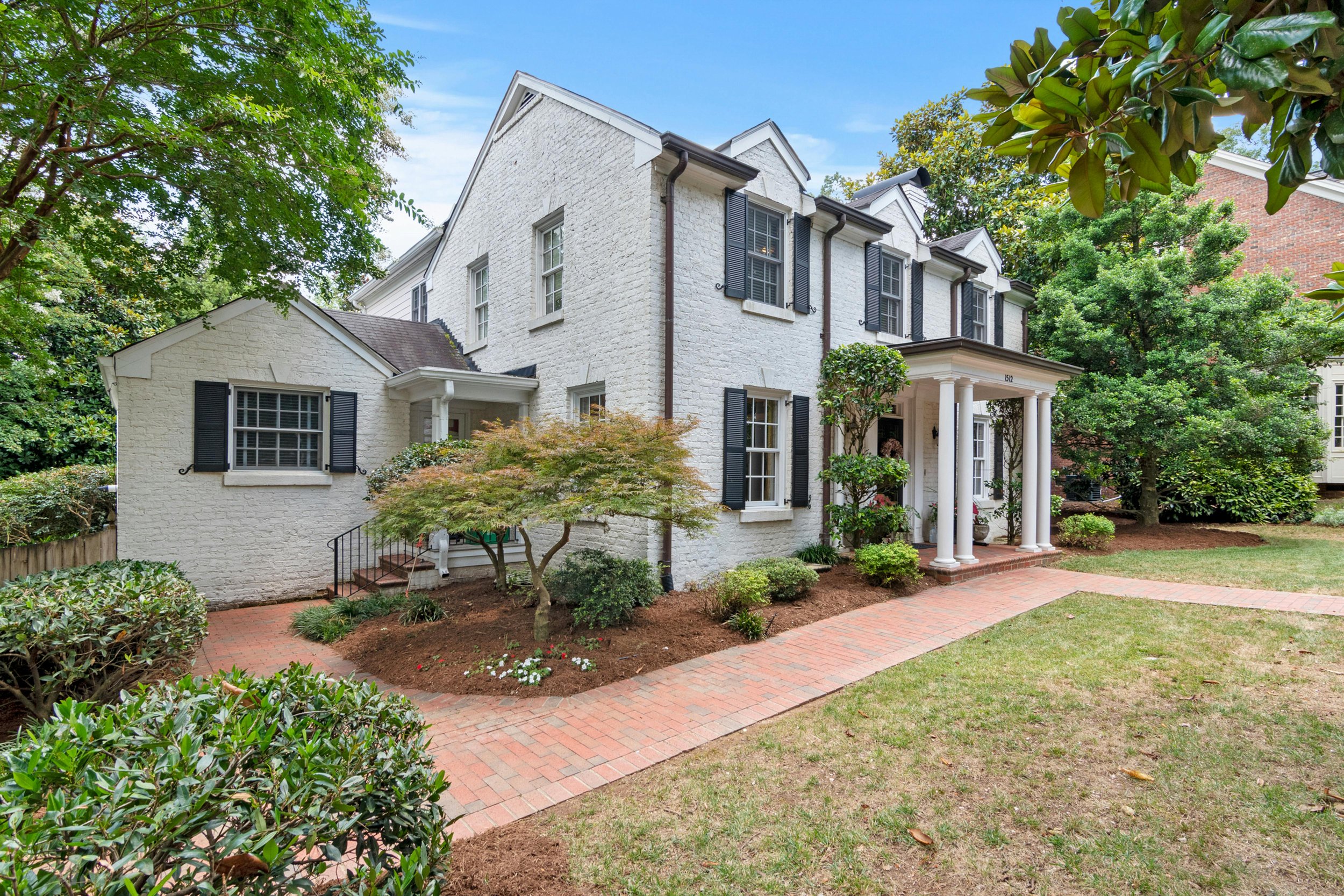 A white brick house with black shutters, a porch with white columns, and a garden with bushes, trees, and a brick pathway.
