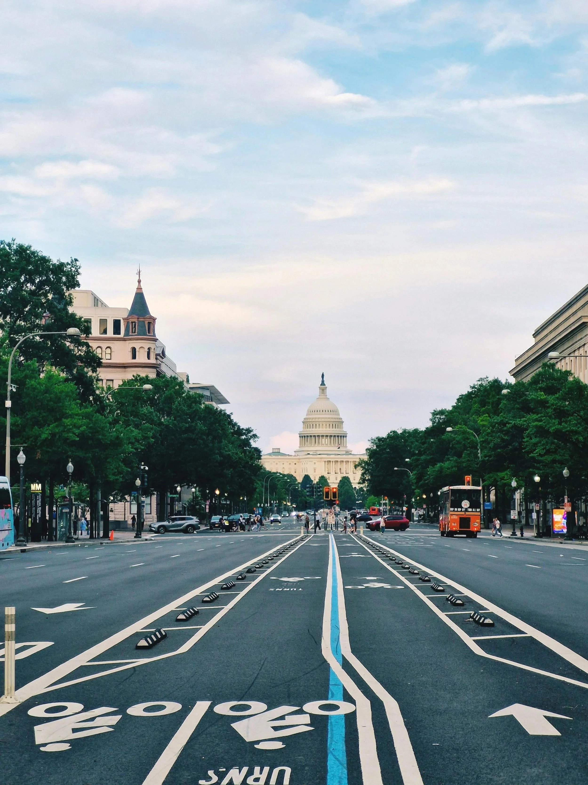A city street leading toward the Capitol building in Washington, D.C. with trees lining the road, traffic signals, a trolley, and pedestrians.