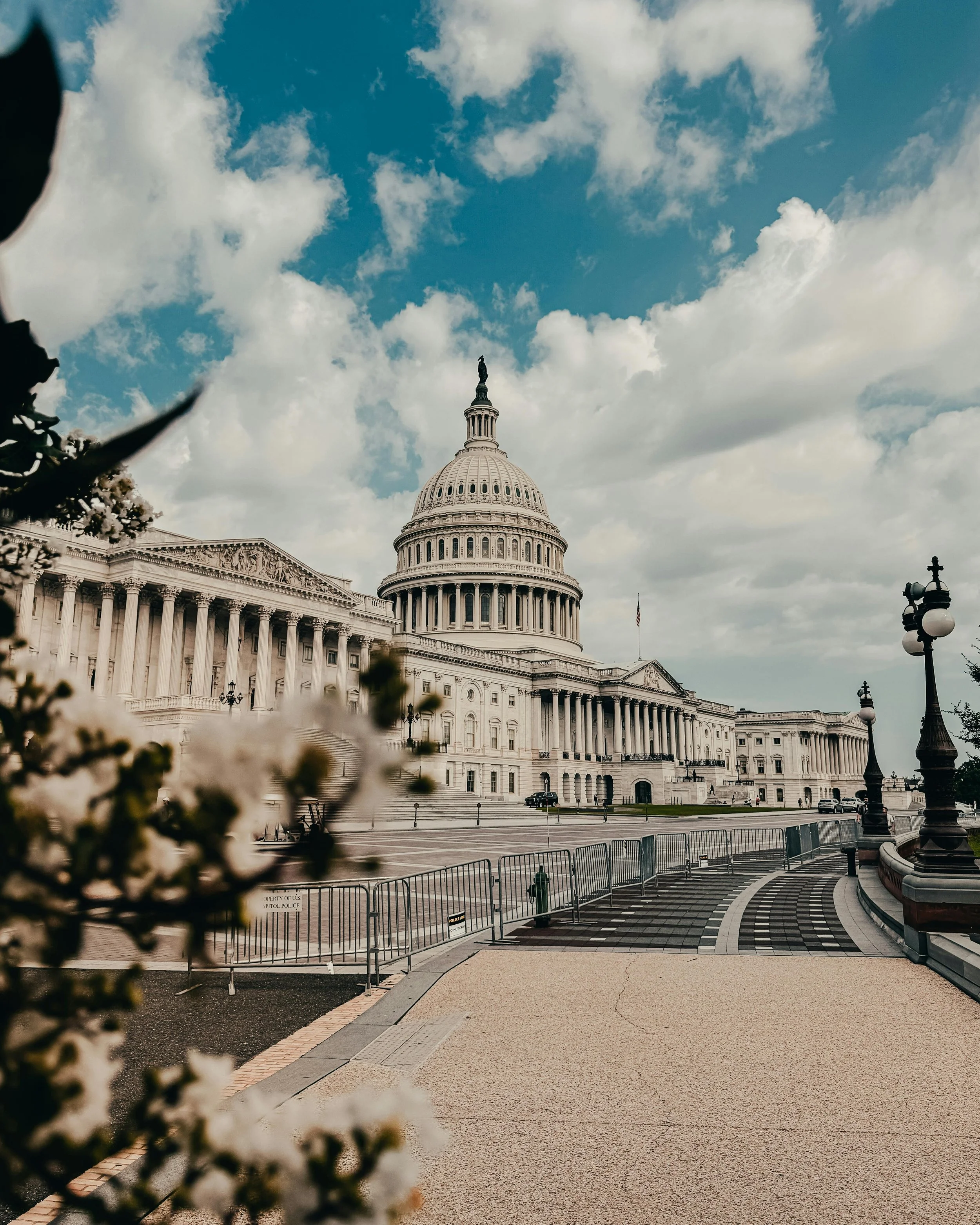View of the United States Capitol building in Washington, D.C., with cloudy sky above, barricades on the street, and a few cars parked near the building.