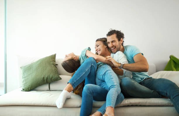 Three young people playfully wrestling and laughing on a couch in a bright living room.