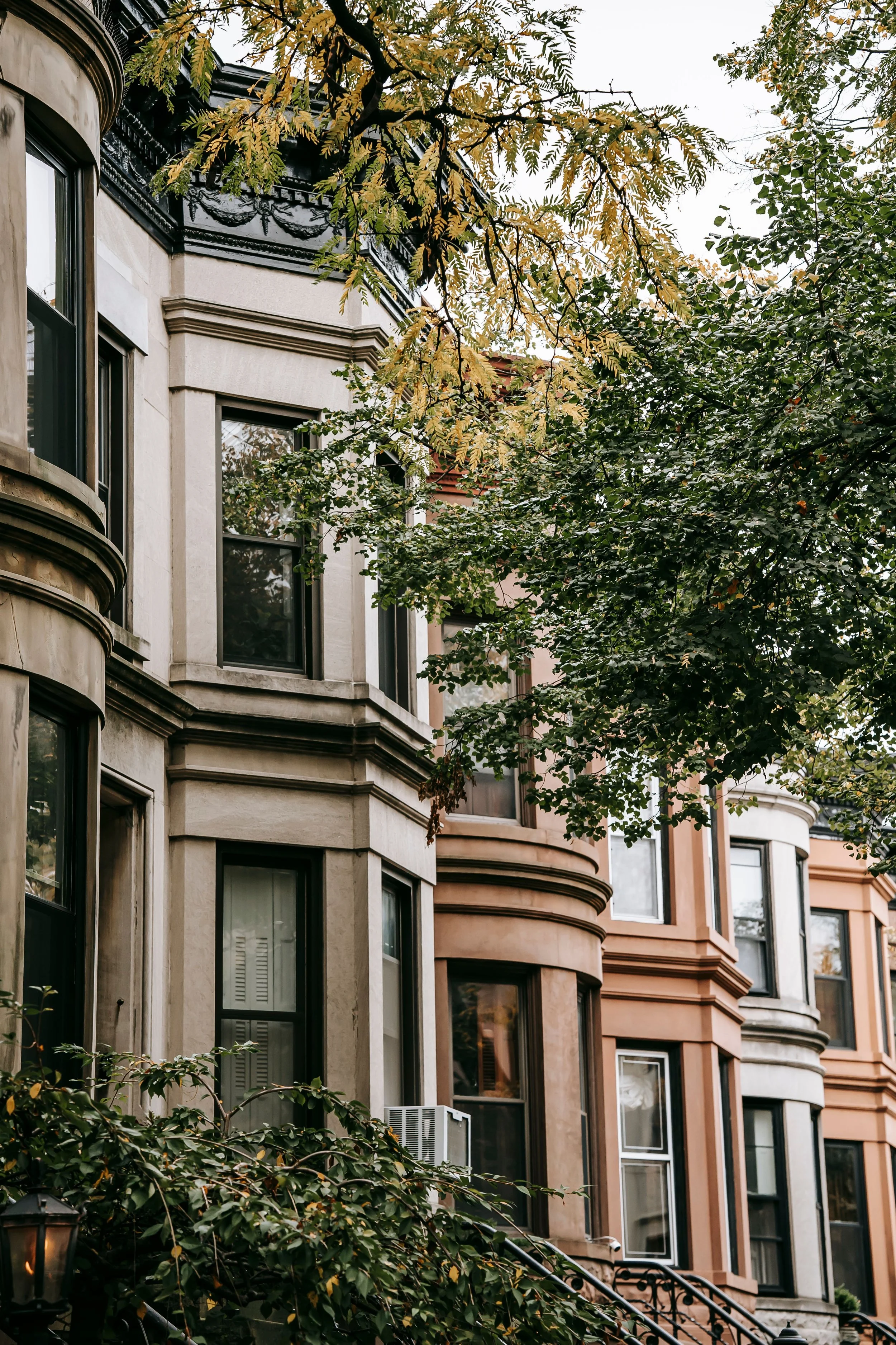 Urban residential street with row of brownstone townhouses, trees with green and yellow leaves, and exterior features like bay windows and stairs in an autumn setting.
