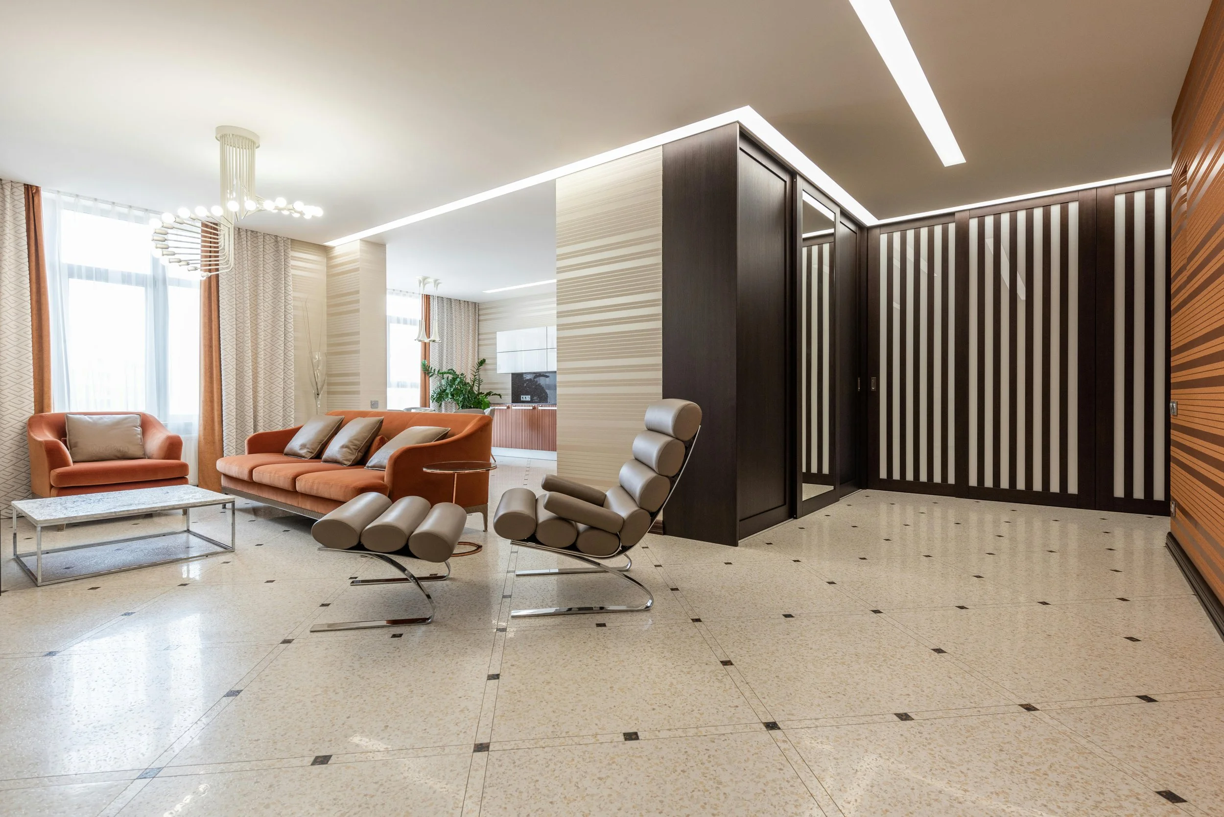 Interior view of a modern hotel lobby with orange couches, olive green and beige armchairs, a marble coffee table, a patterned floor, and large windows with curtains, featuring a hanging chandelier and ceiling lighting.