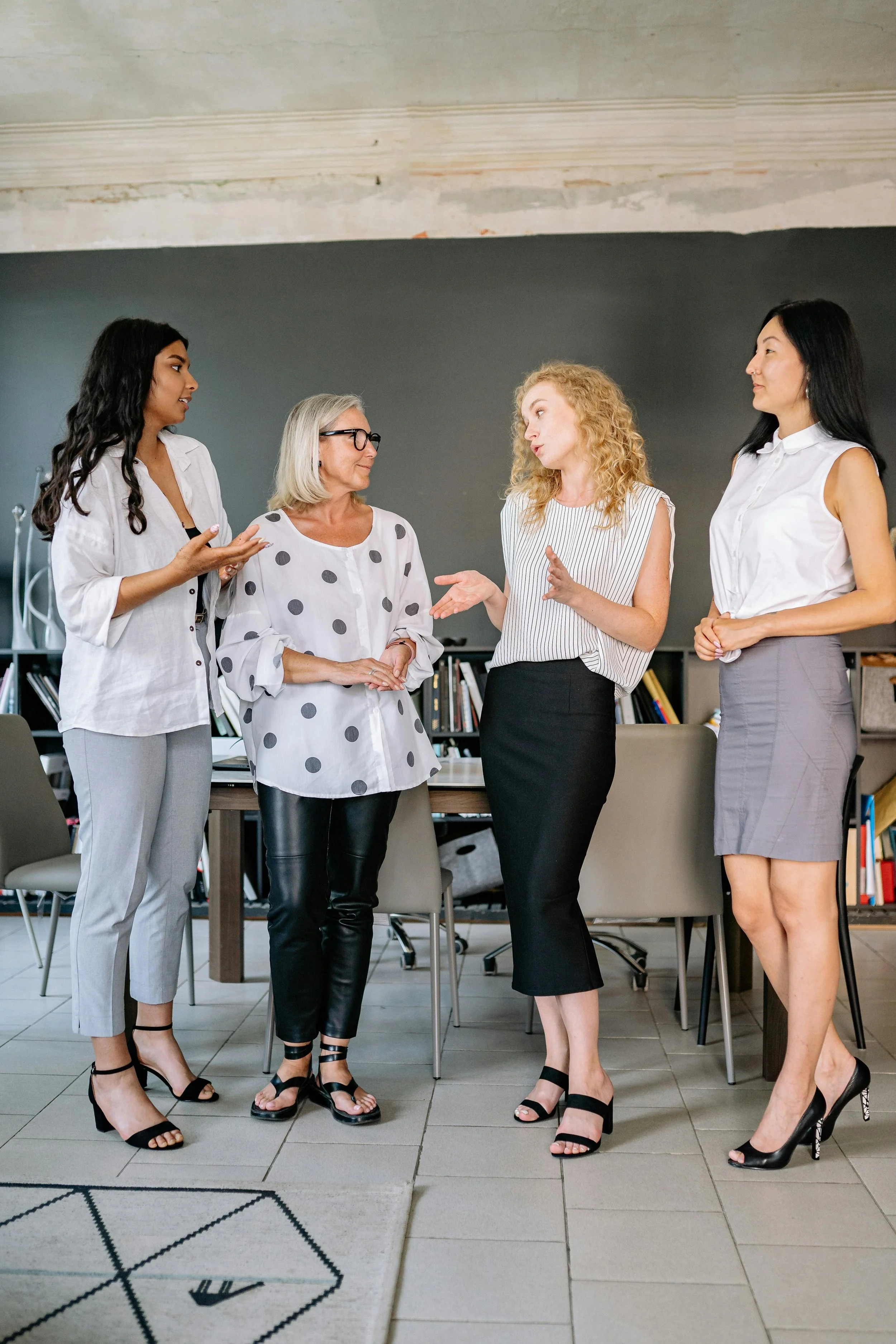 Five women engaged in conversation, standing in a modern office with bookshelves and a gray wall in the background. They are dressed in business casual attire, wearing high heels.