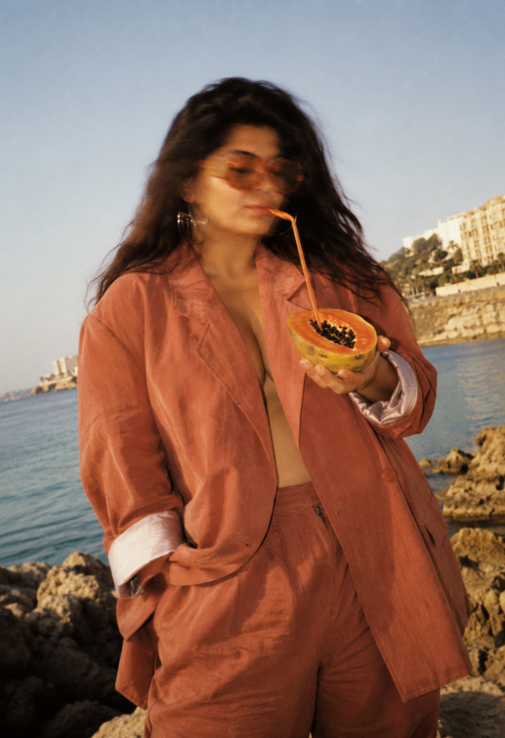 woman in peach suit holding halved papaya at 
rocky coastline during sunset, editorial 
campaign image for Storytotellers