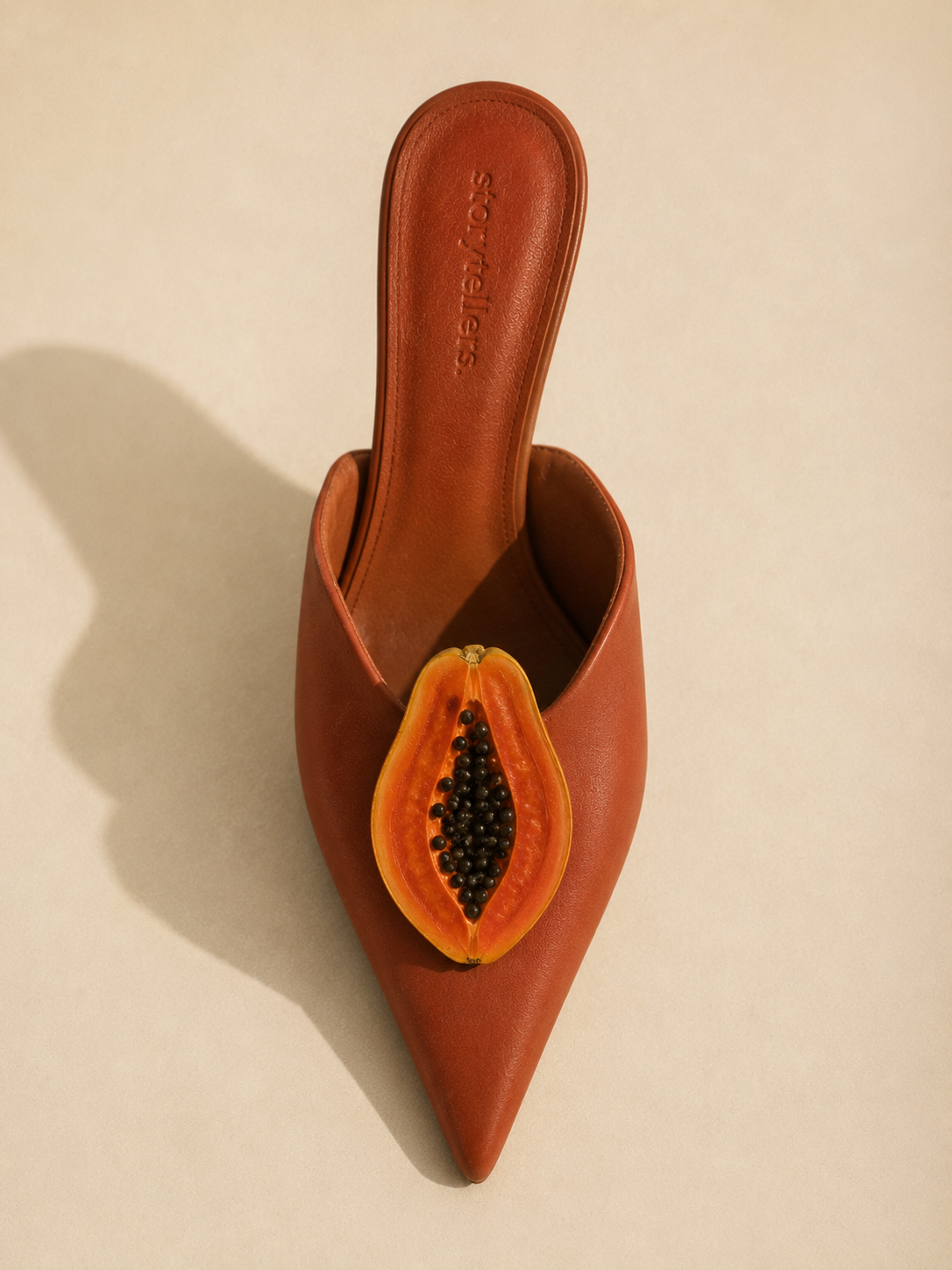 brown leather mule shoe with halved papaya 
revealing black seeds, editorial still life 
for Storytotellers creative consultancy
