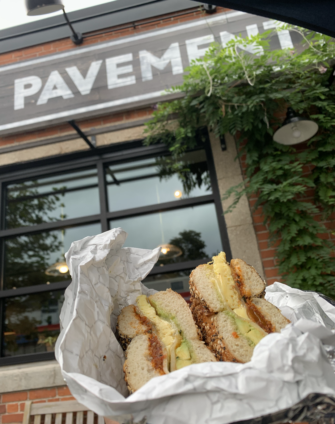 A close-up of a sandwich with avocado, egg, and other ingredients wrapped in parchment paper, held outside in front of a building with a sign that reads 'PAVEMENT' and a window with trees reflected in it.
