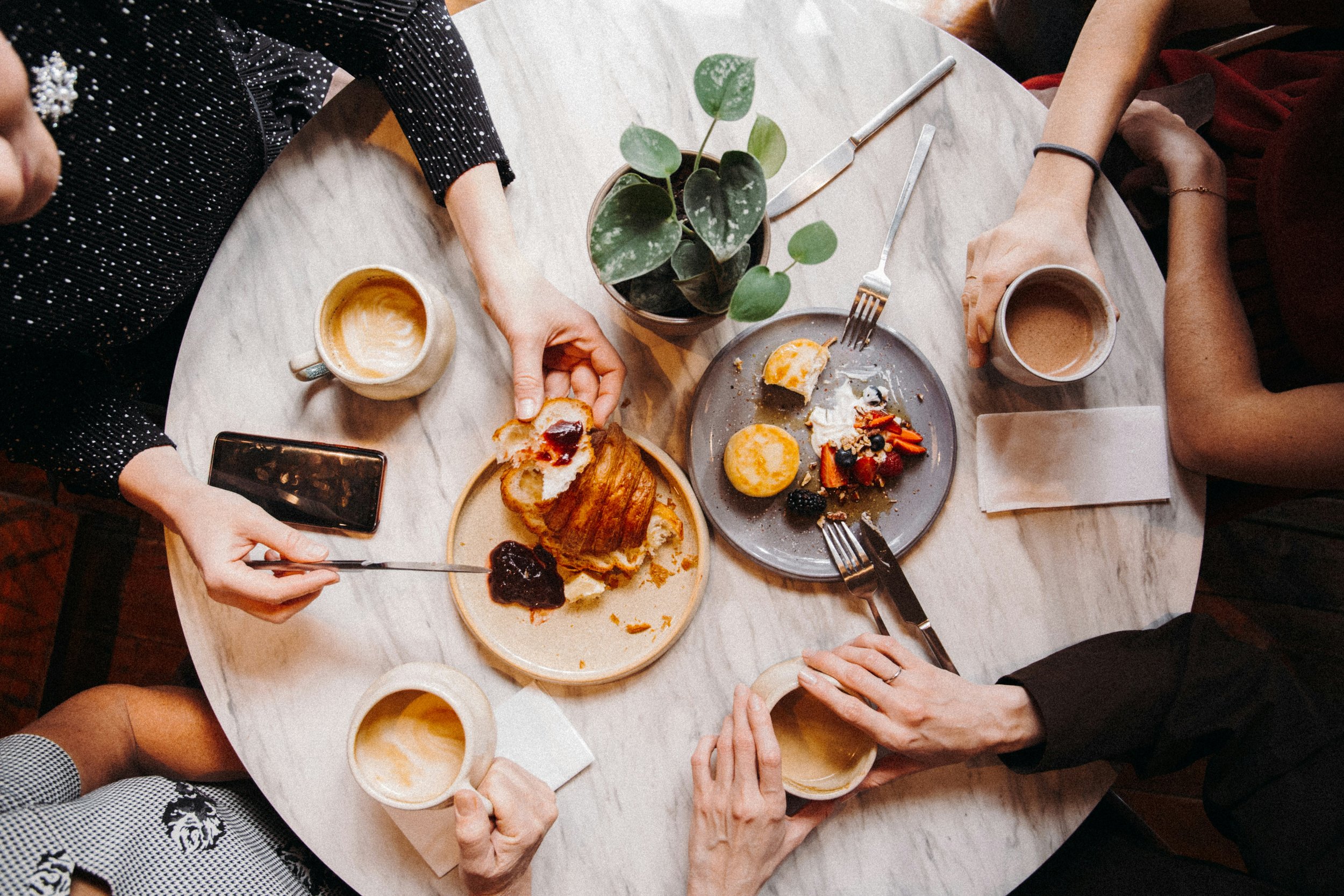 A group of people sitting around a white table with various plates of food, cups of coffee or tea, a potted plant, and a smartphone. The image is taken from above, showing hands reaching for food and holding cups.