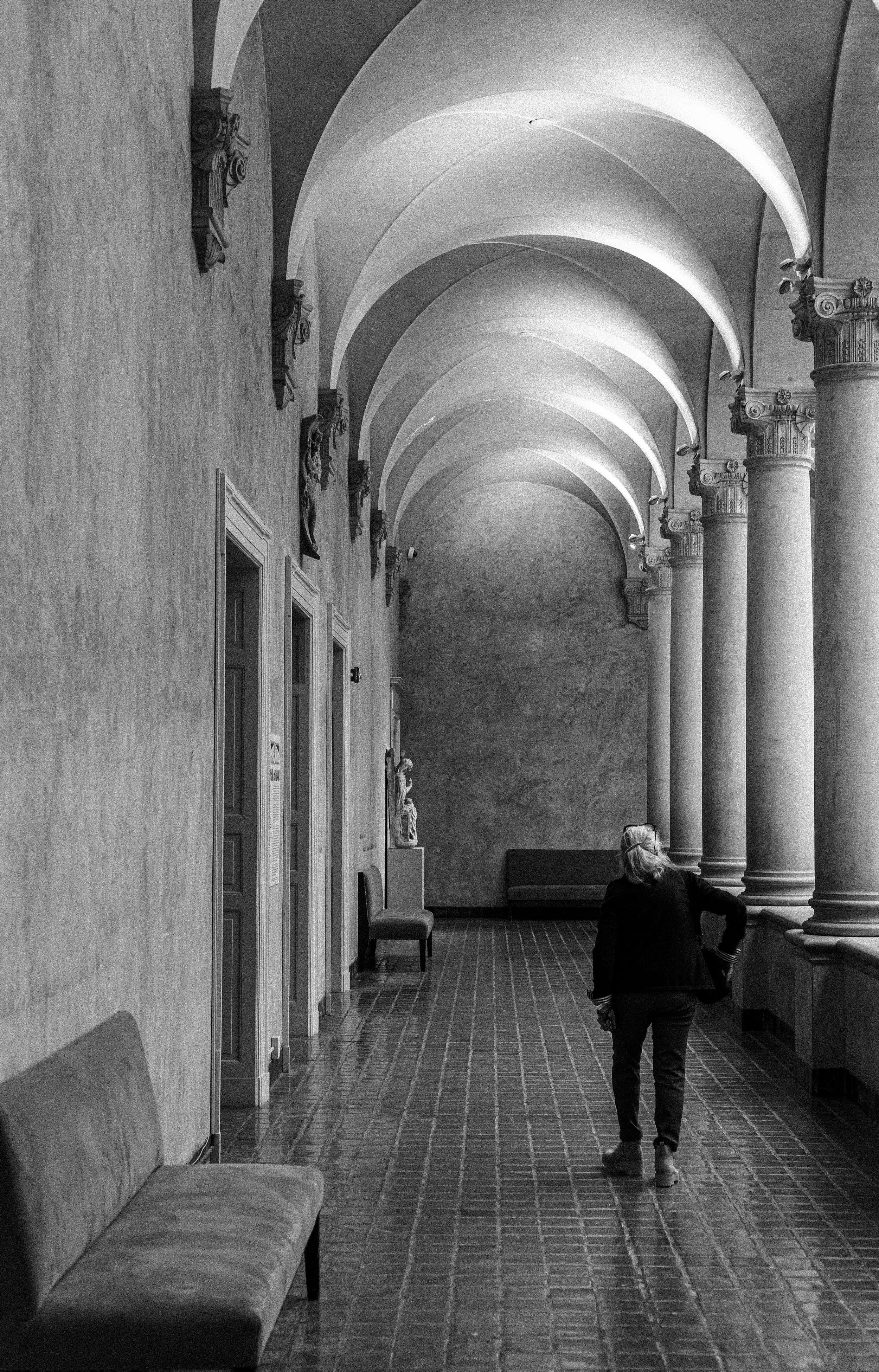 A person with gray hair walking down a corridor with arches, columns, and decorative sculptures on the wall, in black and white.