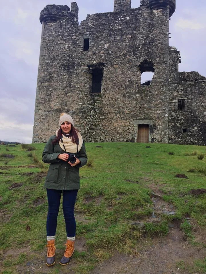 A woman stands on grassy ground in front of a stone castle, holding a camera, dressed in a jacket, scarf, hat, and orange boots.