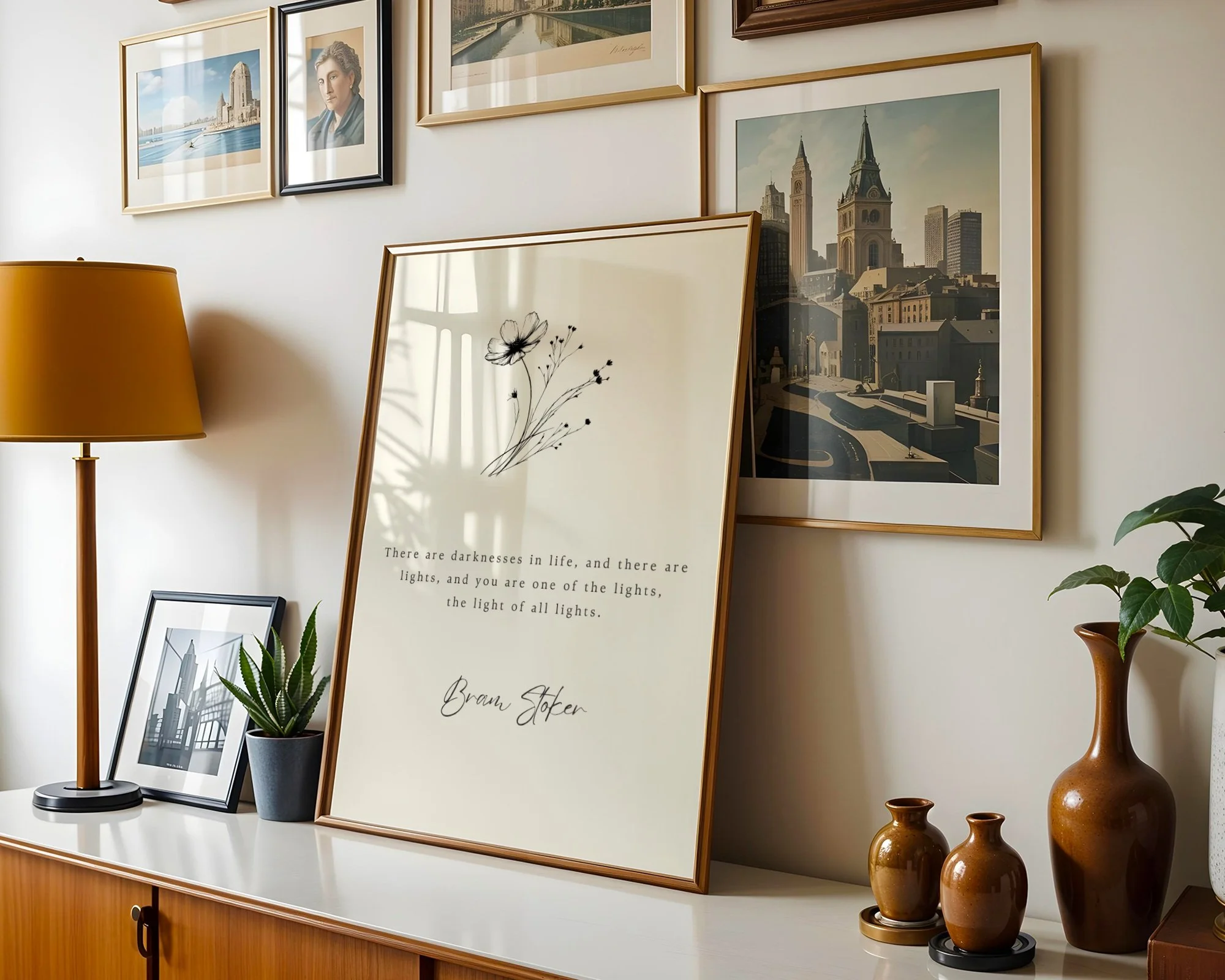 A gallery wall with framed pictures and prints, including cityscapes, a woman's portrait, and a nature scene. A floor lamp with a mustard yellow shade stands to the left, and various potted plants and decorative vases are on a wooden sideboard below the artwork. One print has a quote by Bram Stoker.