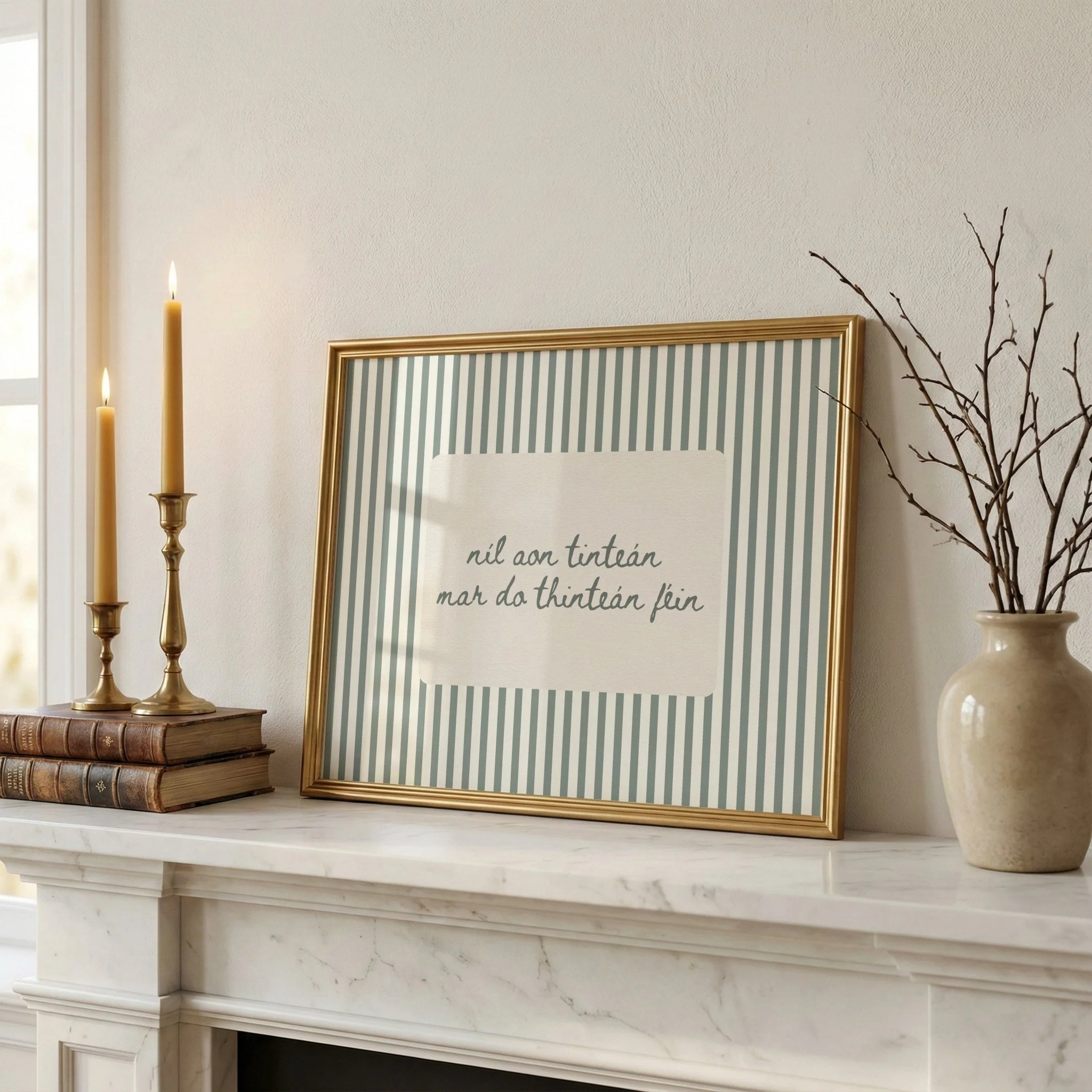 A white marble fireplace mantle decorated with three candles on the left, an empty picture frame with blue and white stripes, a stack of vintage books, and a ceramic vase with dried branches.
