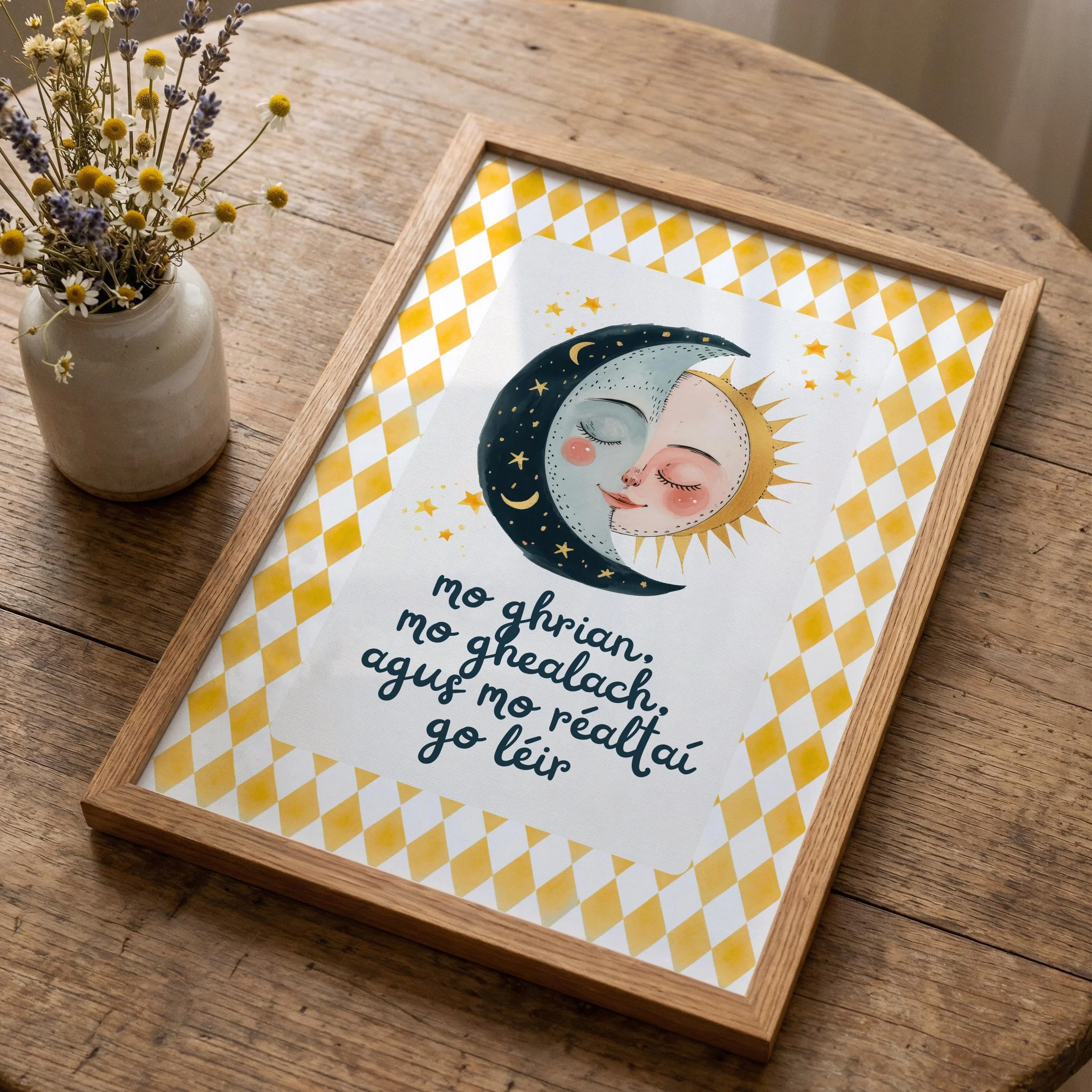 A framed print featuring a celestial design with a crescent moon, sun, stars, and clouds, placed on a rustic wooden table next to a beige vase with wildflowers.