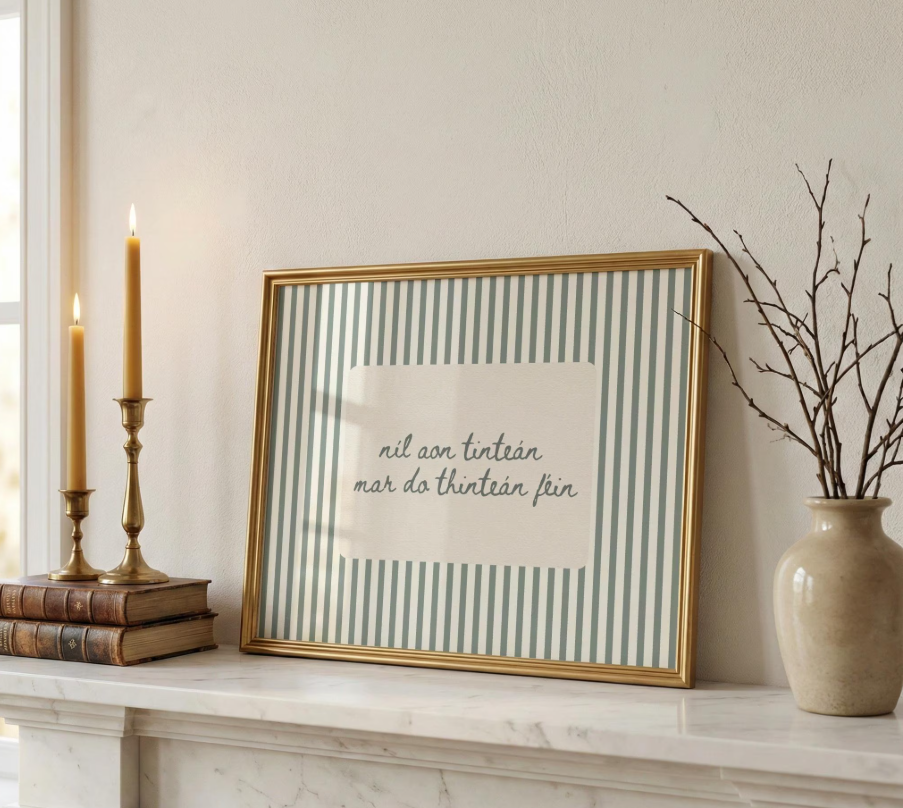 A framed print with striped background and Irish phrase, placed on a white marble mantelpiece next to a beige vase with branches and two lit candles in brass holders, with a window in the background.