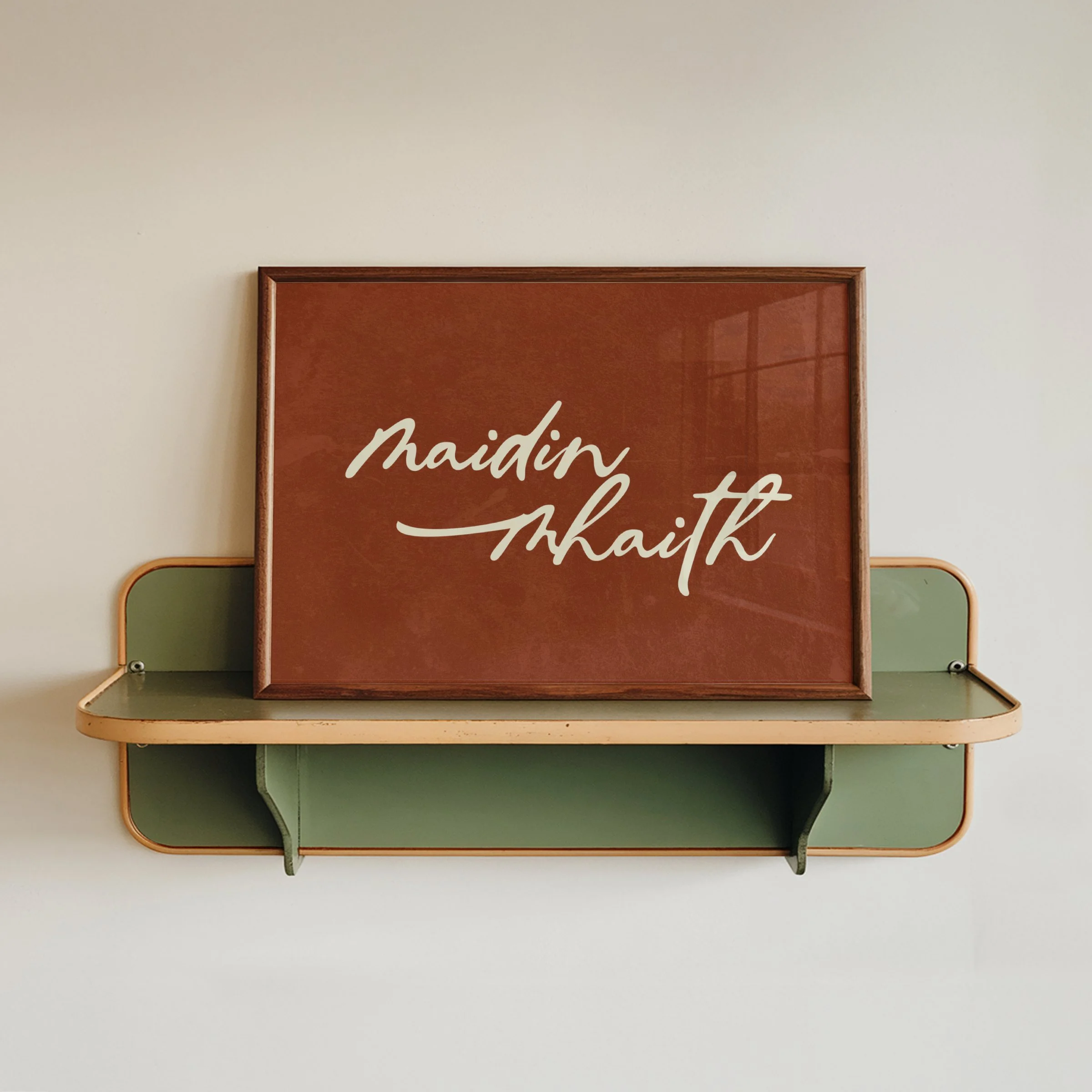 Decorative framed wall art with text 'maidin mhaith'  meaning good morning in Gaeilge (Irish language) on a beige wall, with a wall-mounted shelf below.
