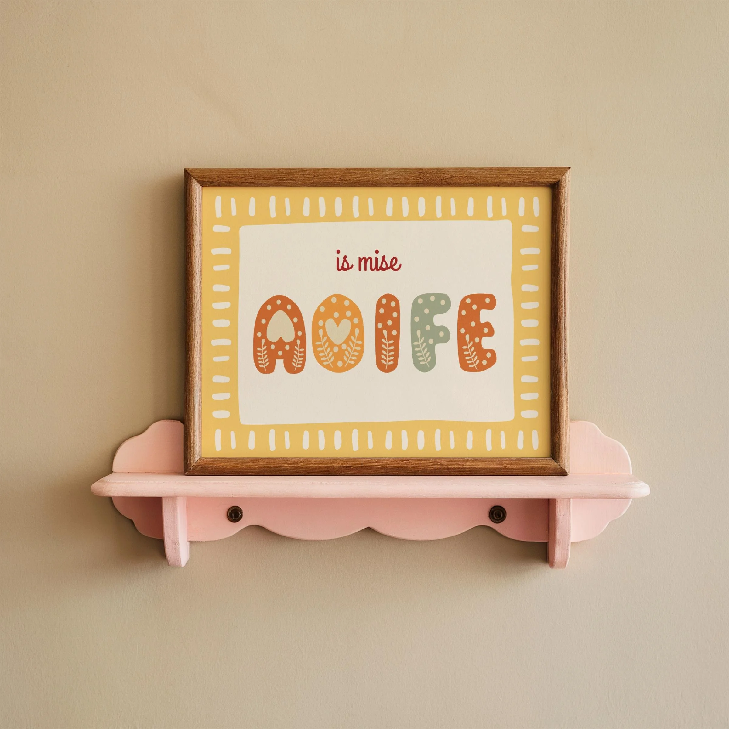 A framed printable with colorful text on a yellow background displayed on a pink shelf against a beige wall.