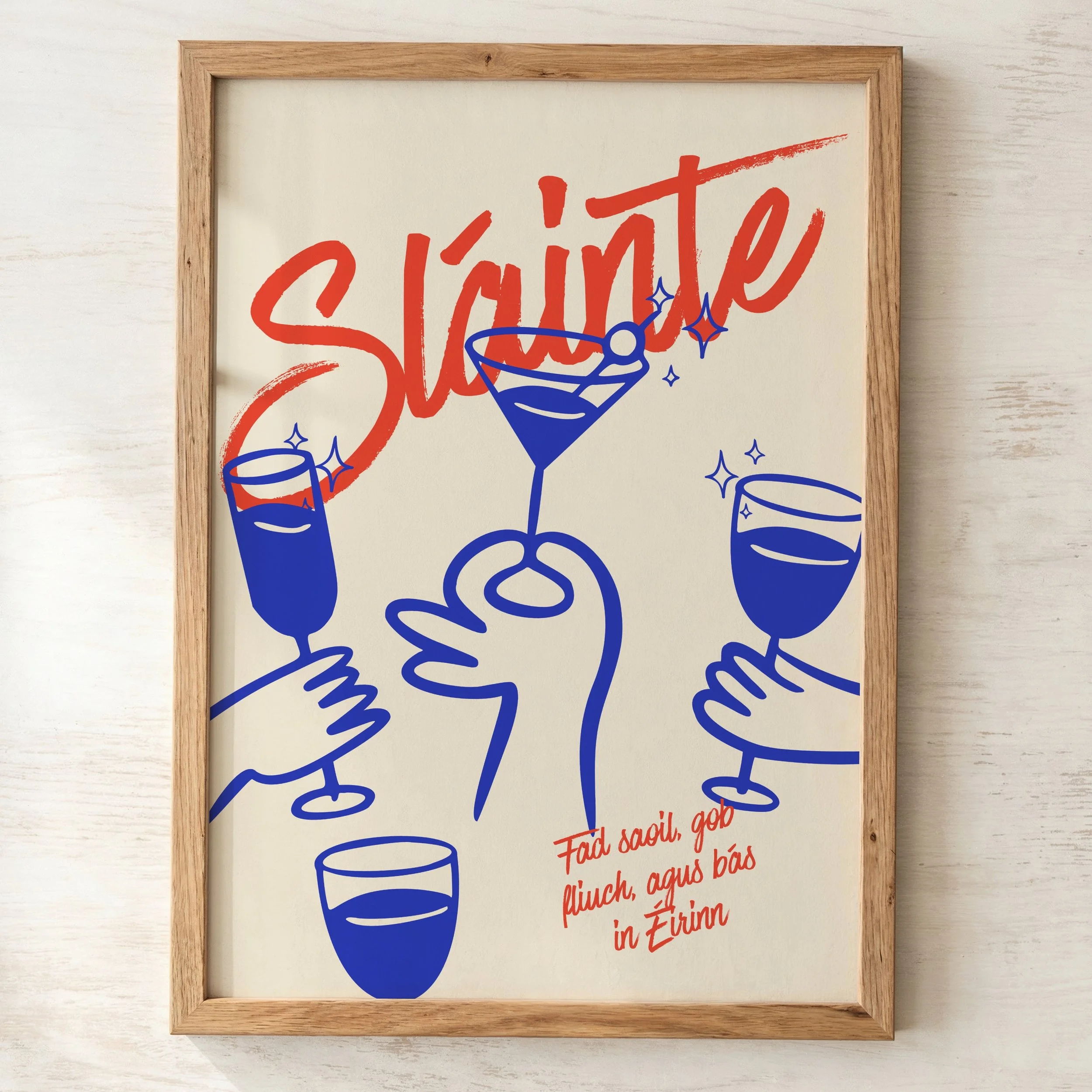 A framed poster with the word "Shine" in large red script, and an illustration of a blue cocktail glass with sparkles and a lemon wedge, on a light wooden surface.