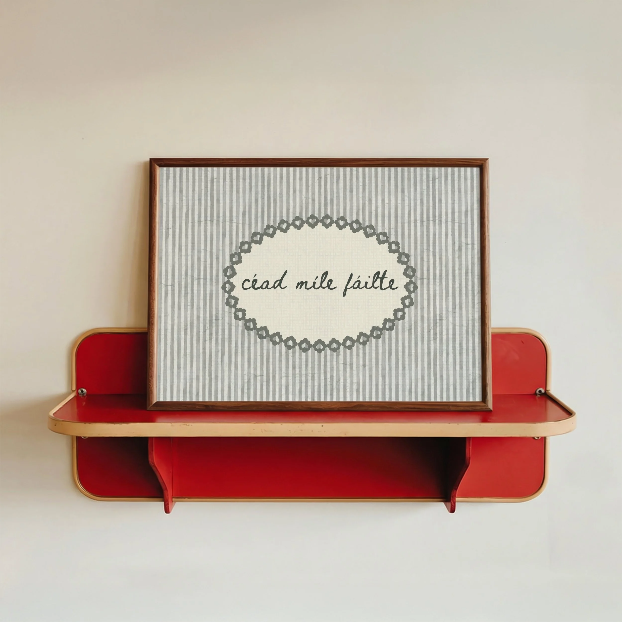 A framed print with a decorative heart-shaped border and the phrase "céad míle fáilte" on a striped background, placed on a red and beige wall-mounted shelf.