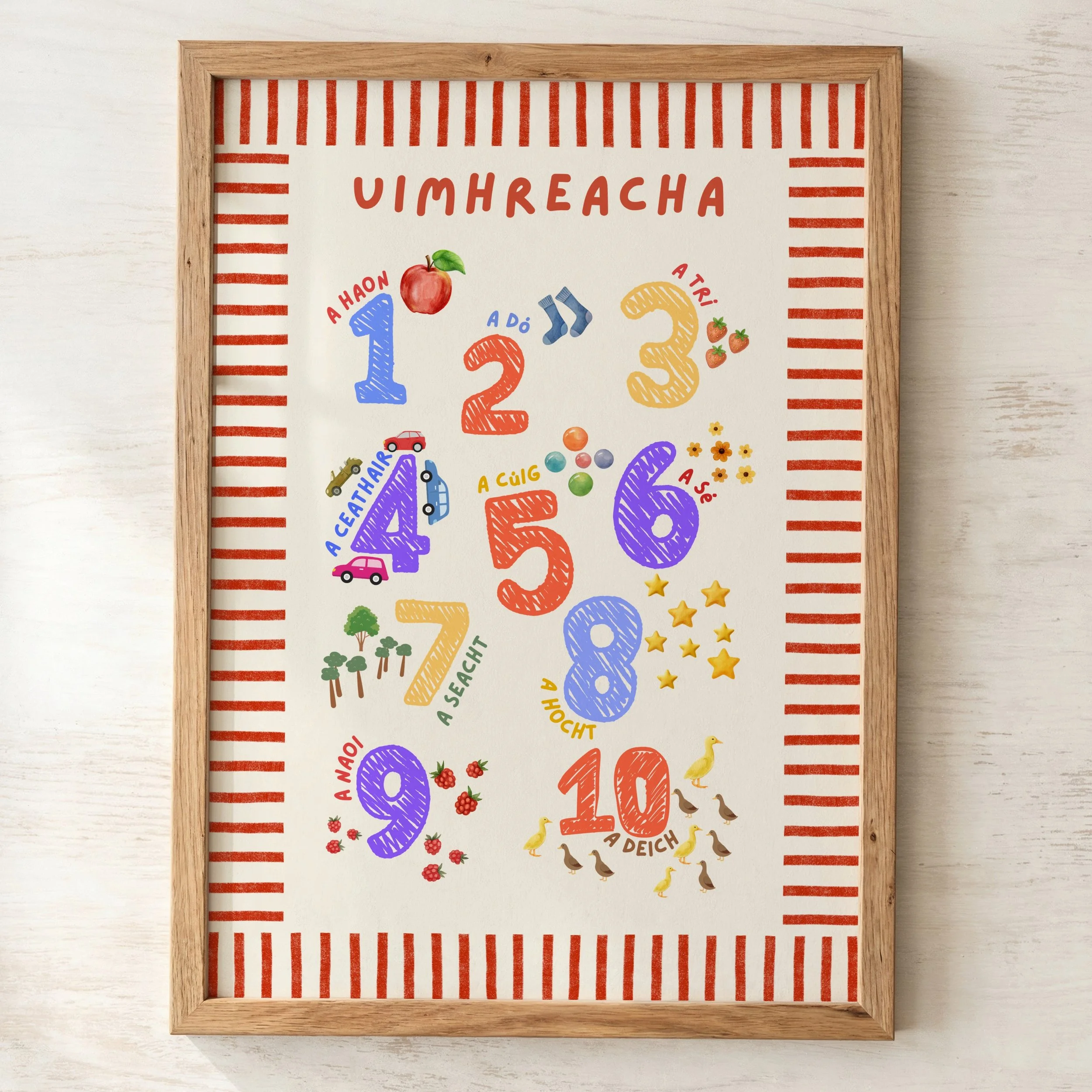 Colorful Irish language educational poster inside a wooden frame, displaying math numbers and words in a playful style, with a striped red border, on a white wall.
