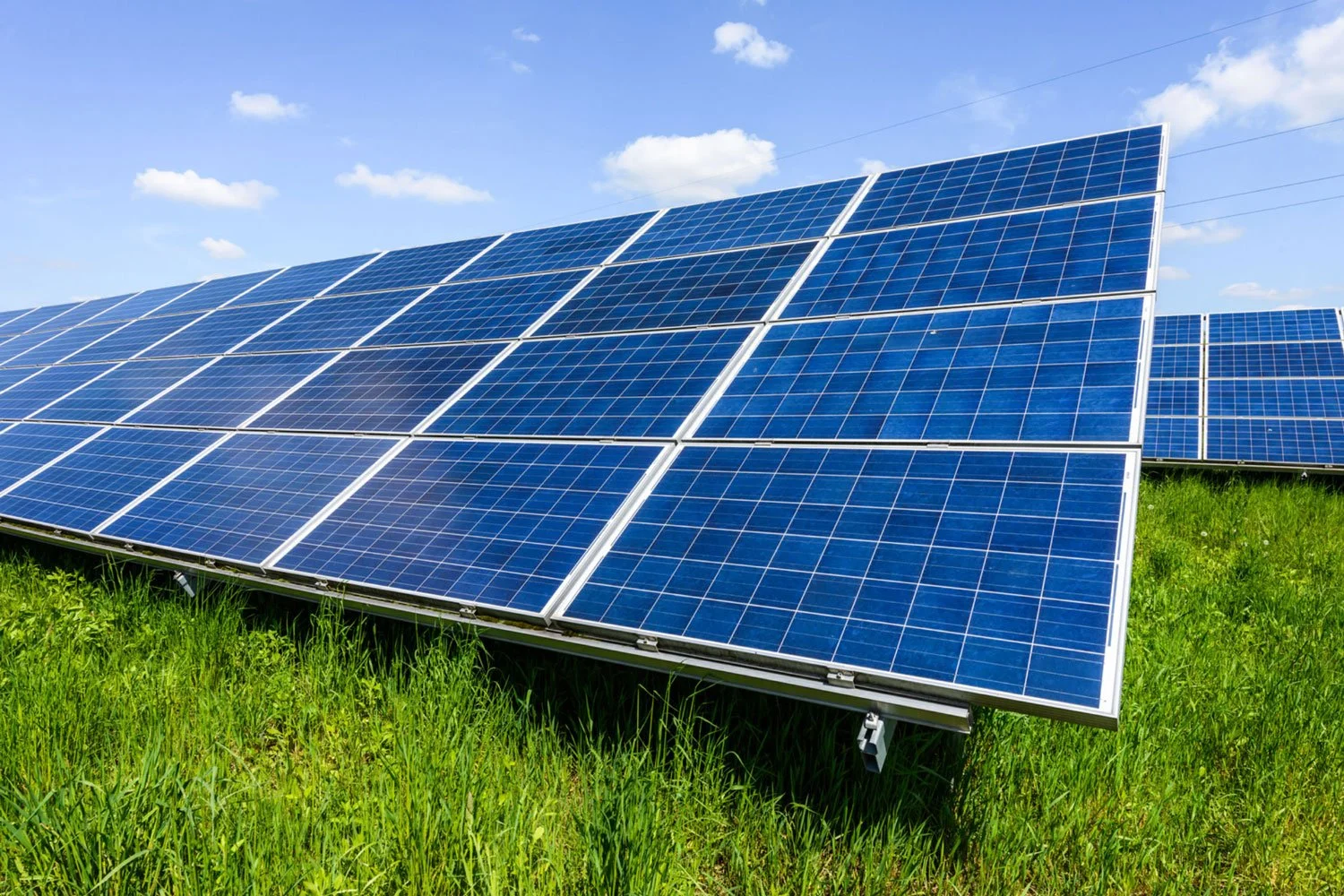 Multiple rows of blue solar panels installed on a grassy field under a partly cloudy sky.