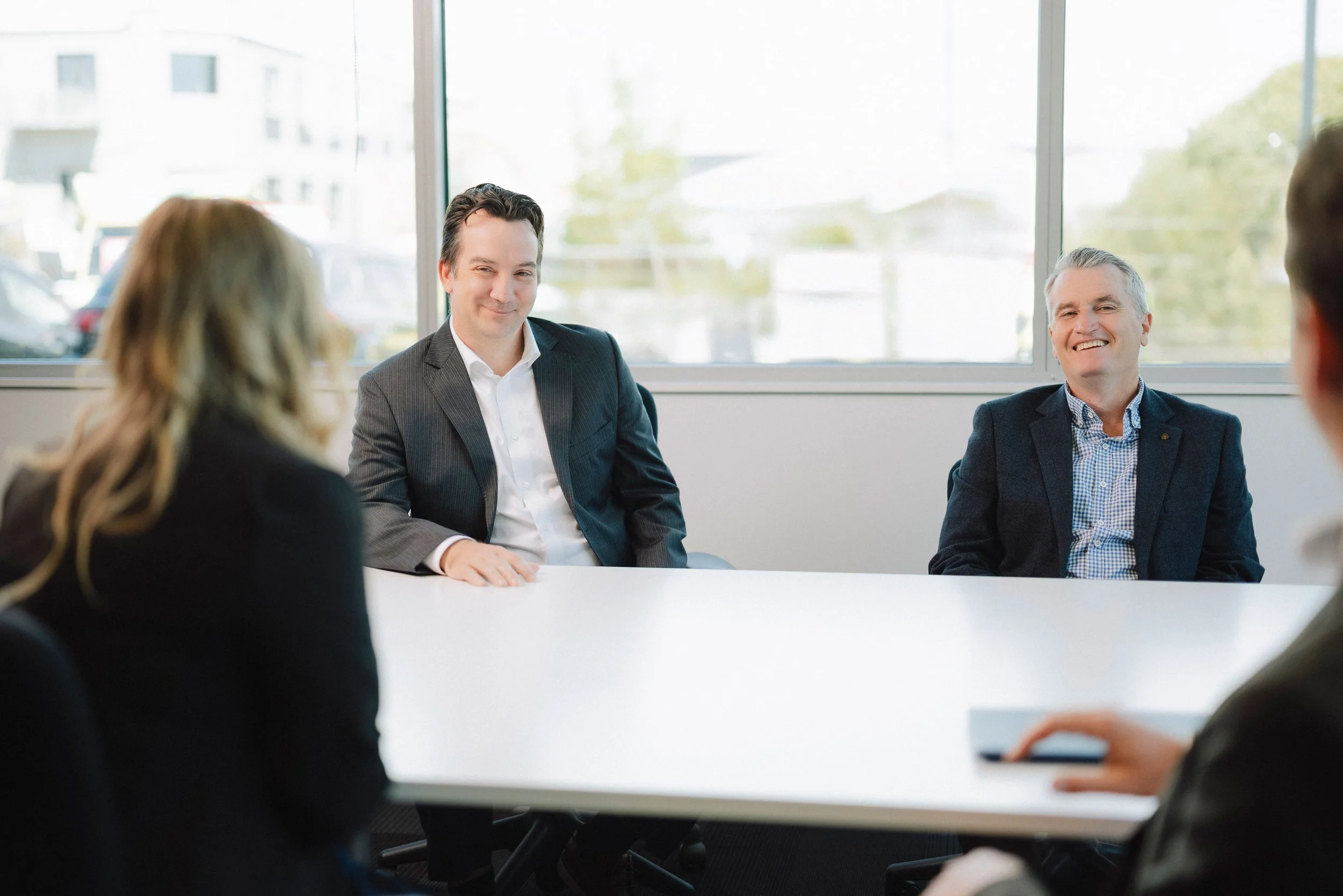 A business meeting with six professionals sitting around a white table in a bright conference room with large windows, some smiling and engaging in conversation.