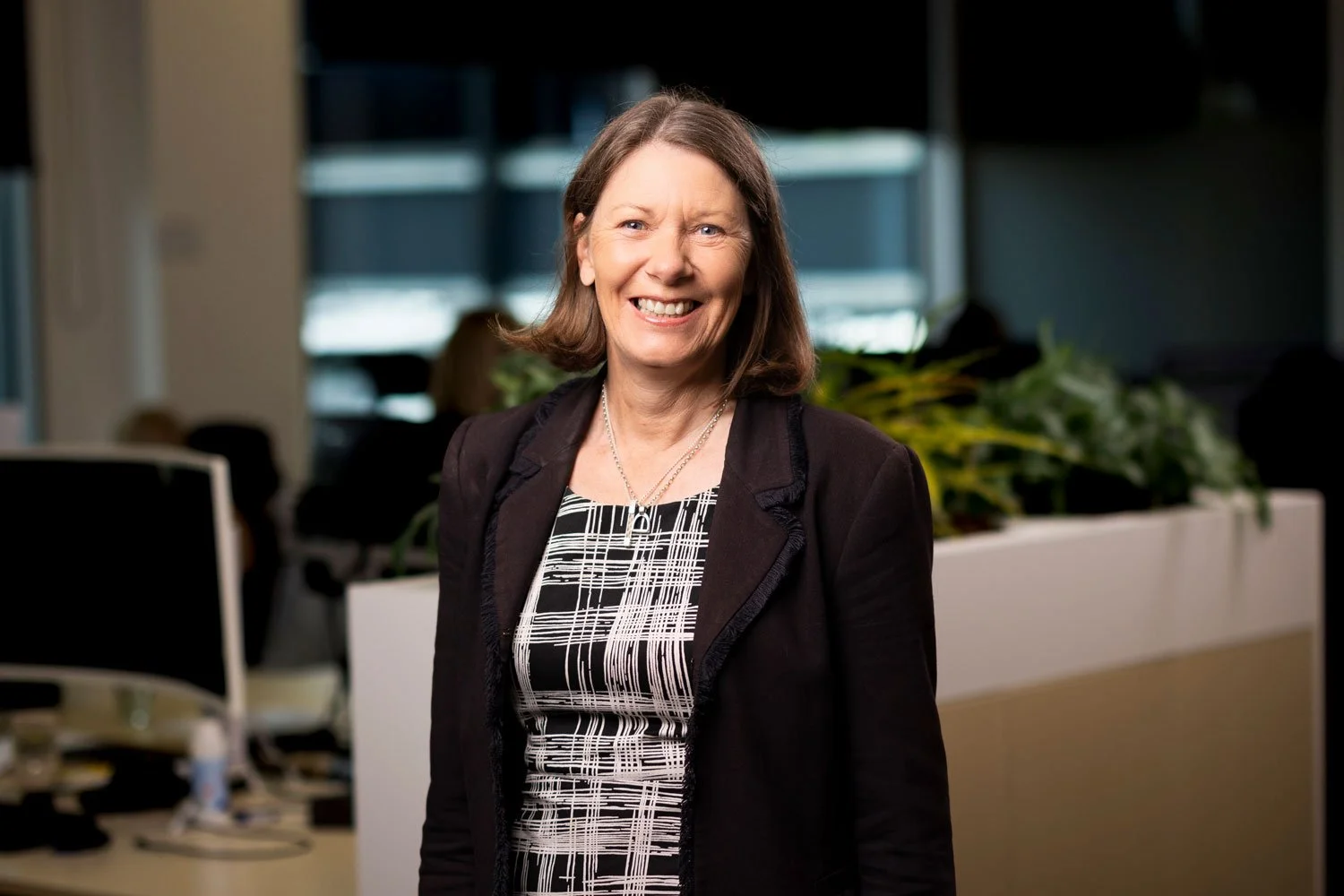 A smiling woman with shoulder-length brown hair wearing a black blazer and patterned dress standing in an office setting with plants and desks visible in the background.