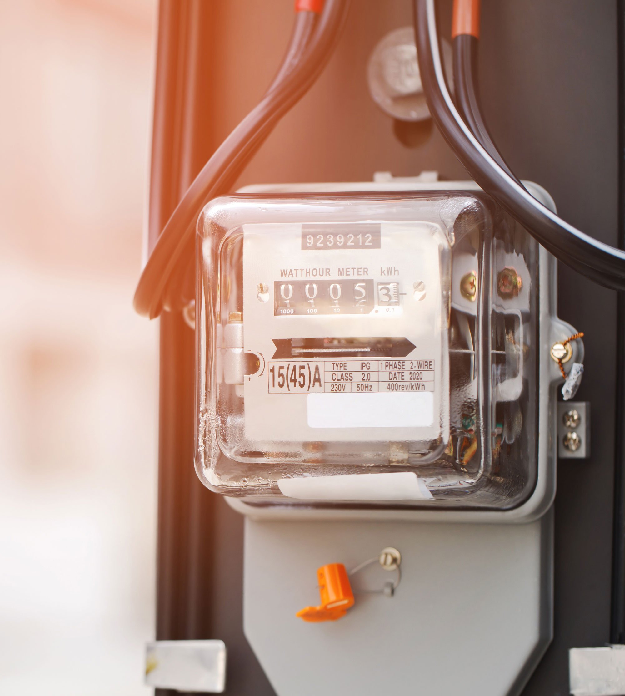 Electric meter displaying 53 kilowatt-hours with orange and black wires connected, mounted on a gray panel.