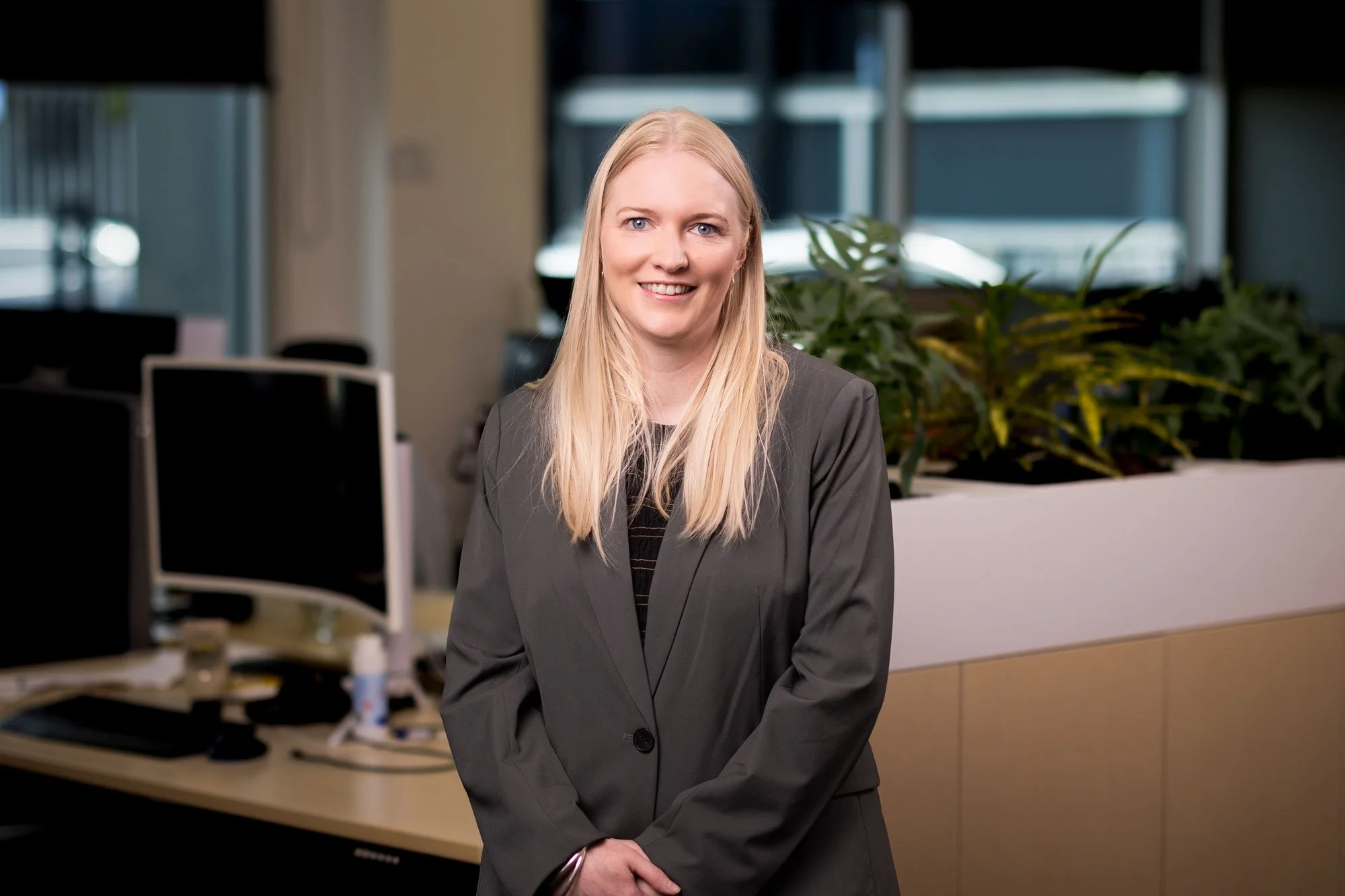 A professional woman with long blonde hair, wearing a gray blazer and smiling, standing in an office with computers, bookshelves, and green plants in the background.