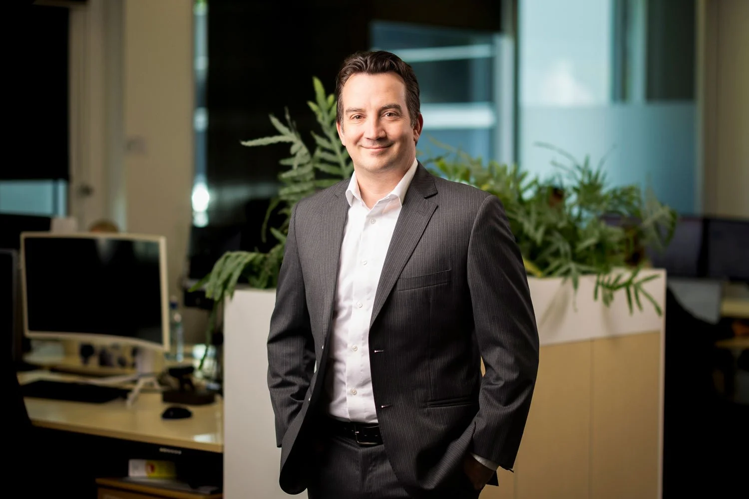 A man in a gray suit with a white shirt smiling in a modern office setting with plants and computer monitors in the background.