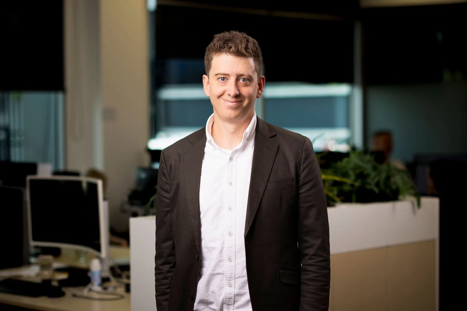 A young man with short brown hair, wearing a white shirt and a black blazer, standing in an office environment with desks, computers, and plants in the background, smiling at the camera.