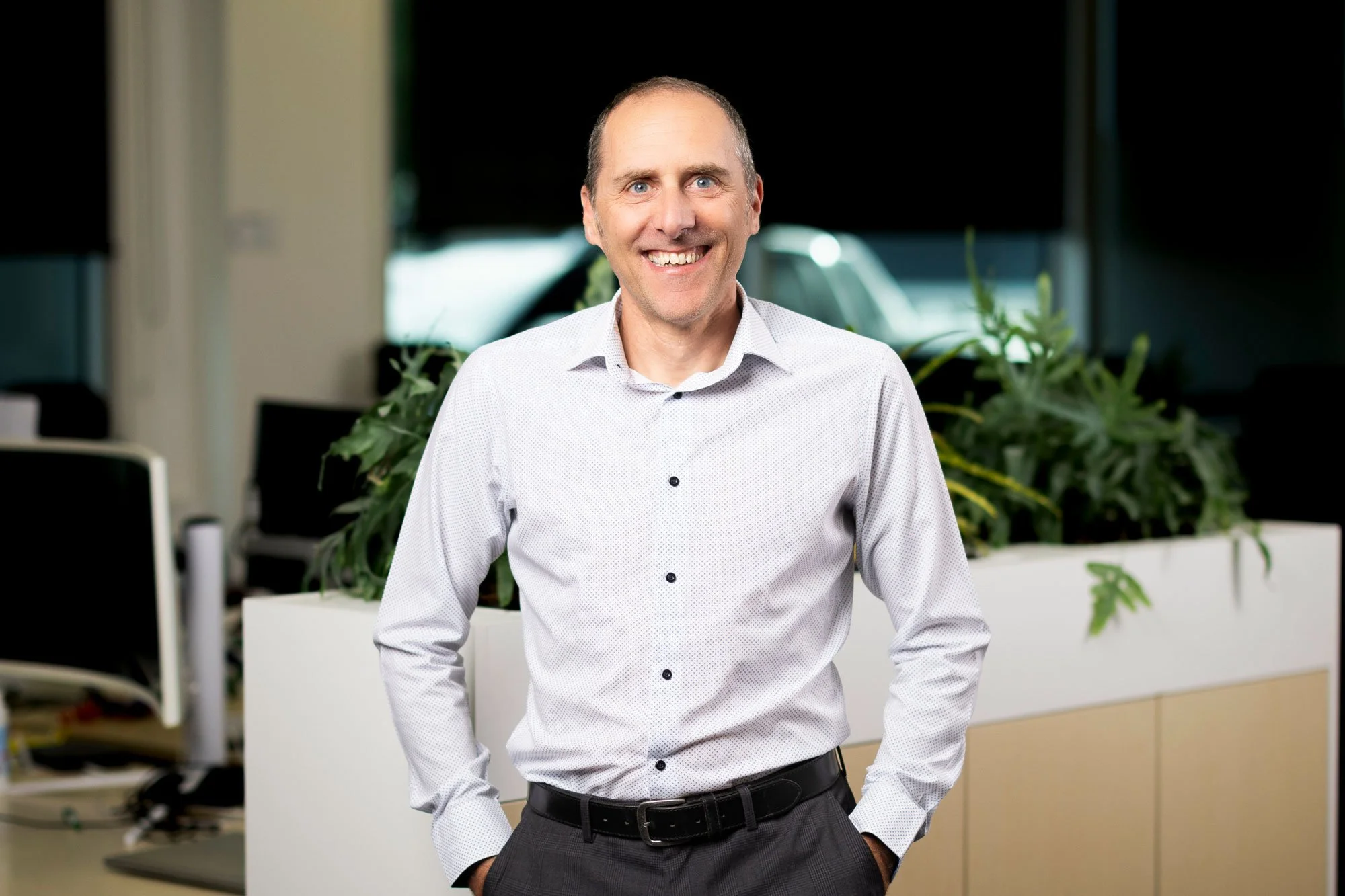Smiling man in a white shirt standing in an office with plants and computer desks in the background.