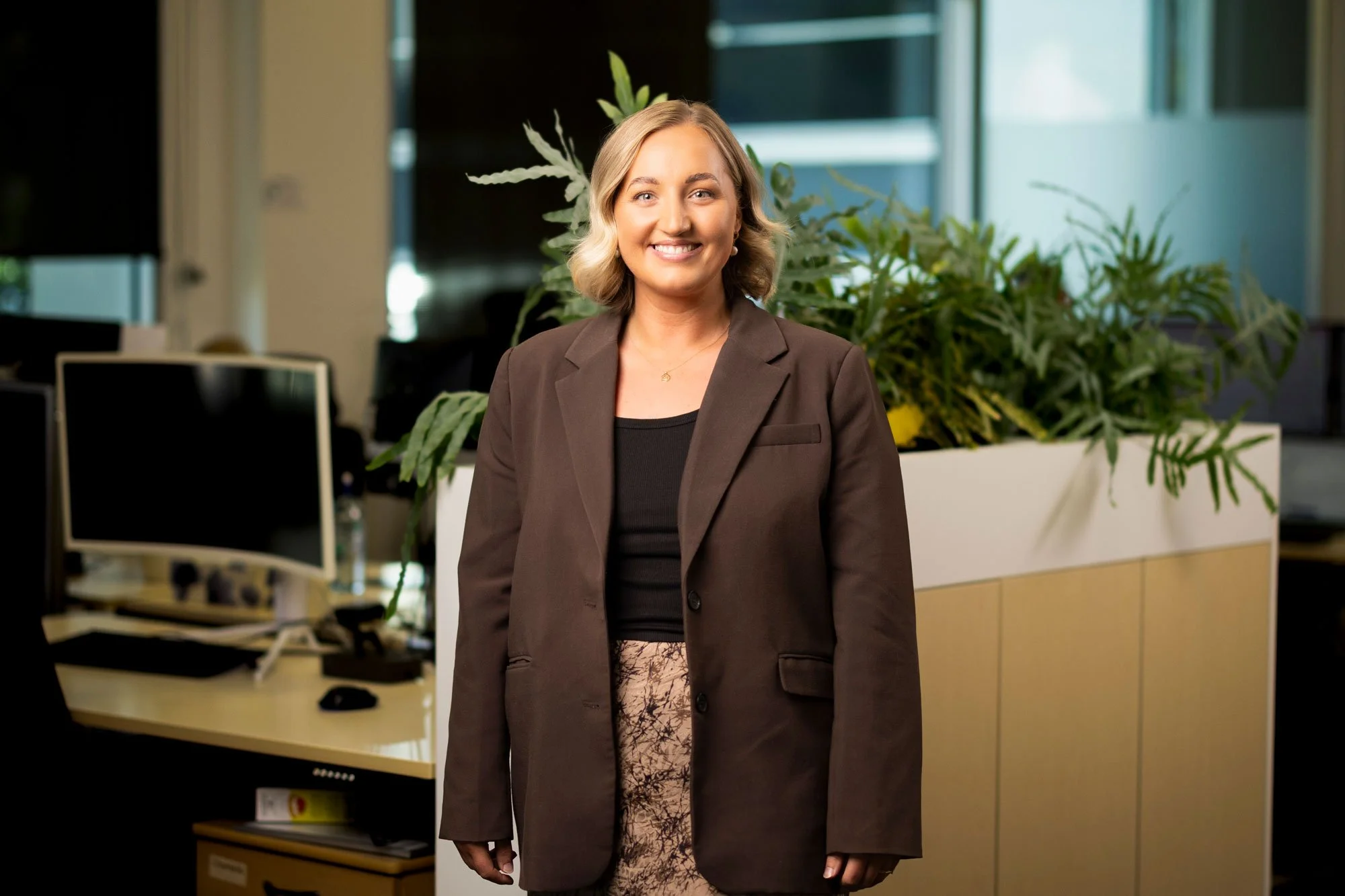 A woman smiling in an office environment with plants and computers in the background.