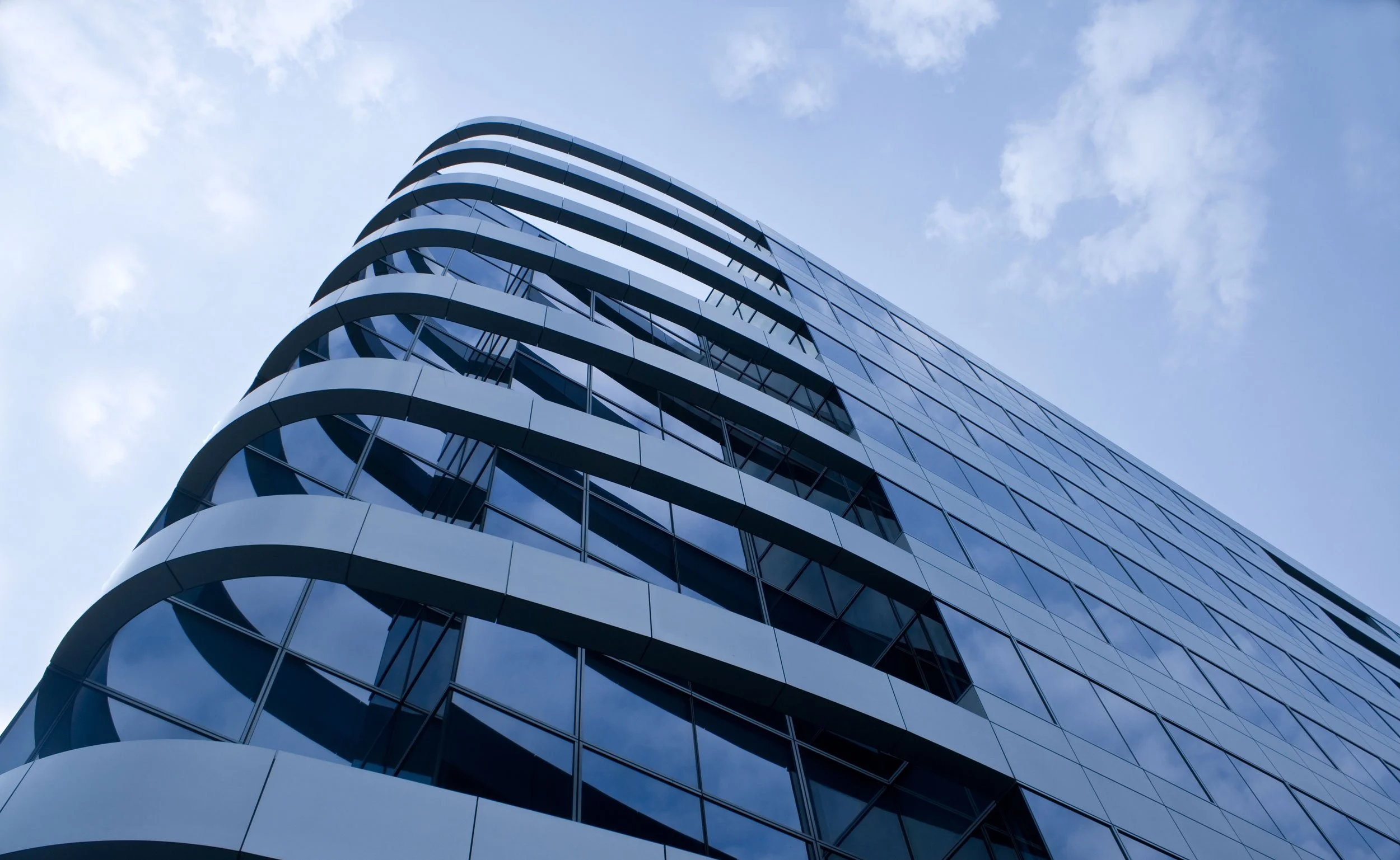 Low-angle view of a modern high-rise building with curved white architectural details and reflective glass windows, against a blue sky with scattered clouds.