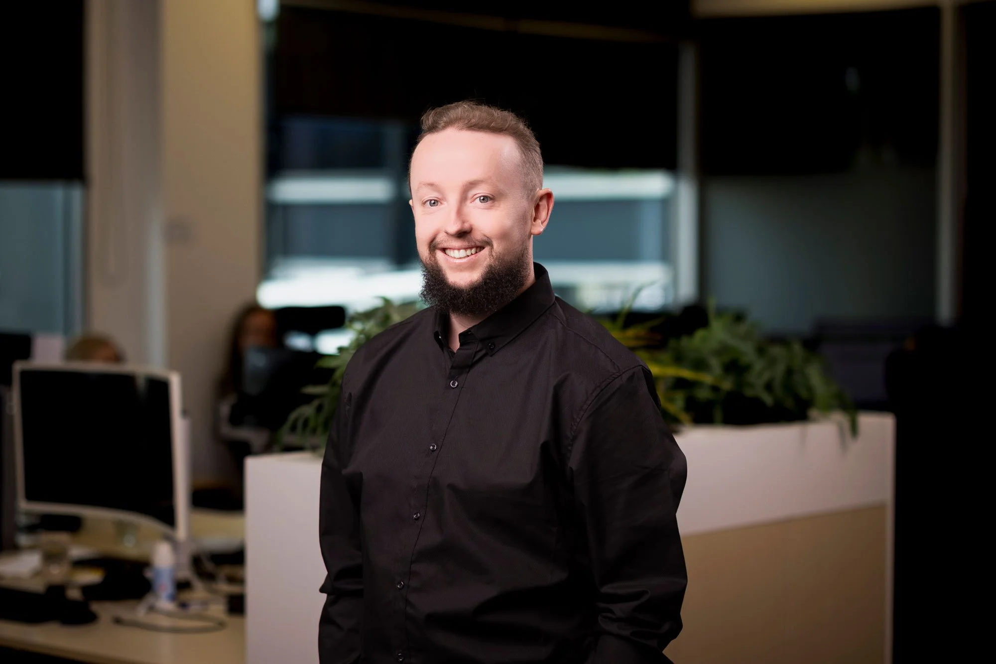 Headshot of a man with short hair, a beard, and a smile, wearing a black button-up shirt, in an office setting with computers and plants.