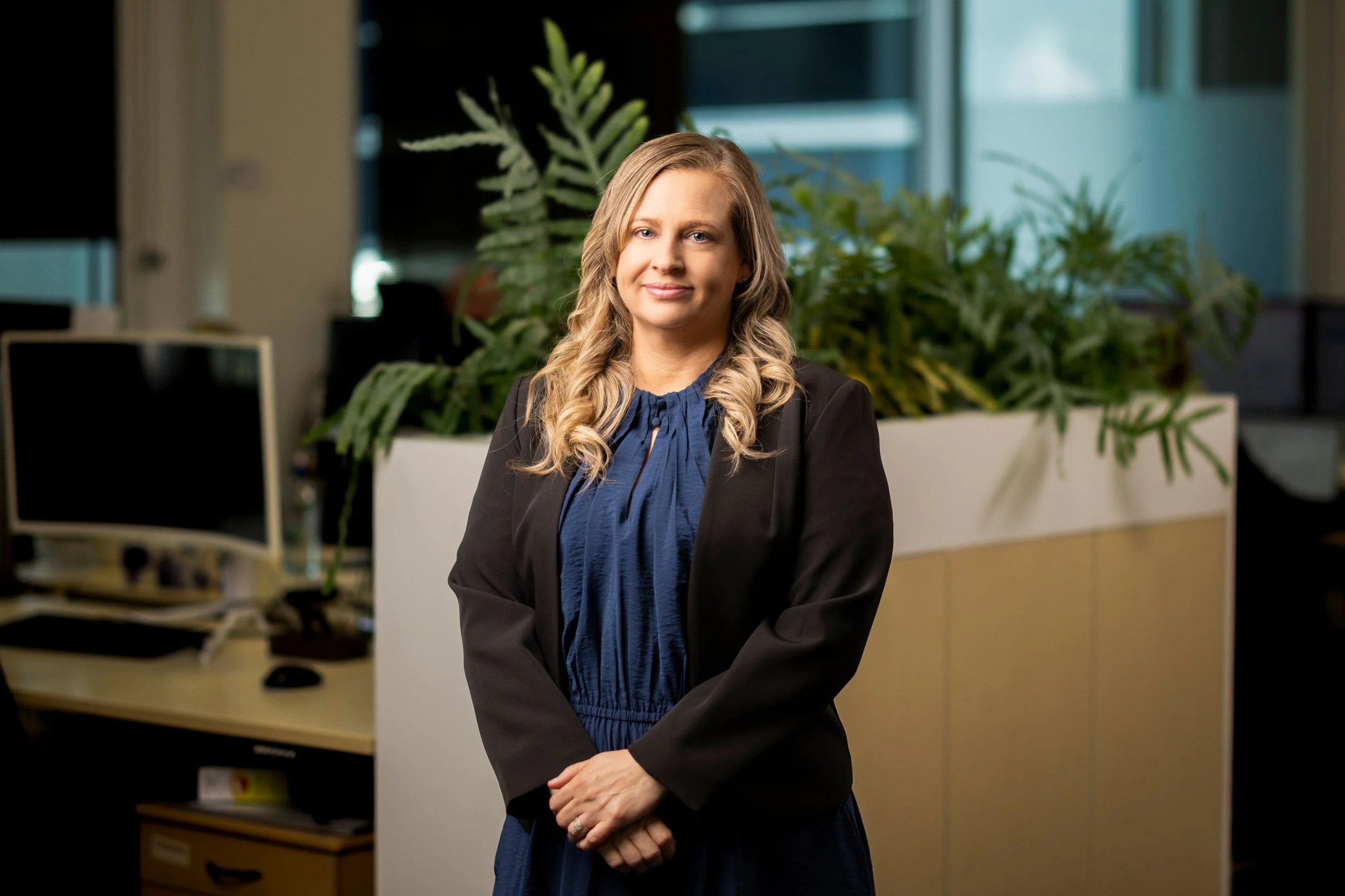 A woman with blond, wavy hair wearing a black blazer over a blue blouse, standing in an office with large windows and green plants in the background.