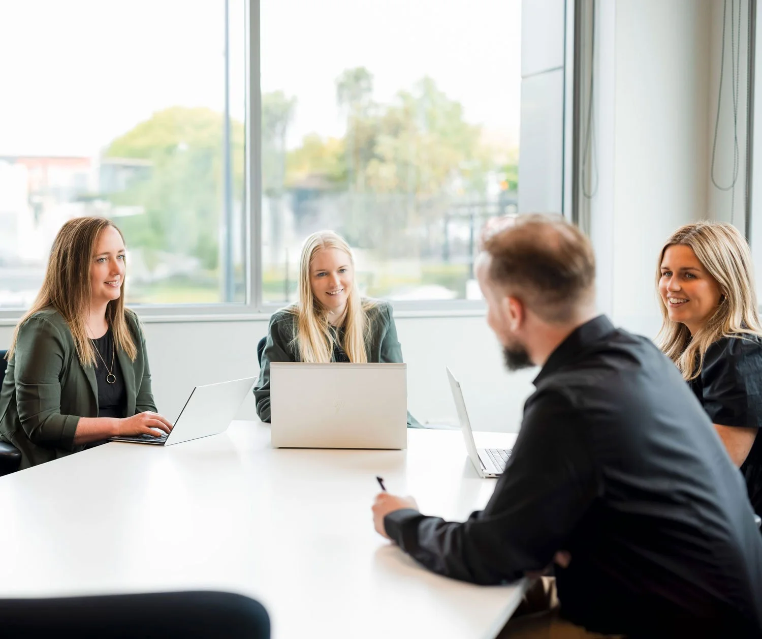 Four young adults in a business meeting in a modern office with large windows, using laptops and a tablet, engaged in conversation and smiling.