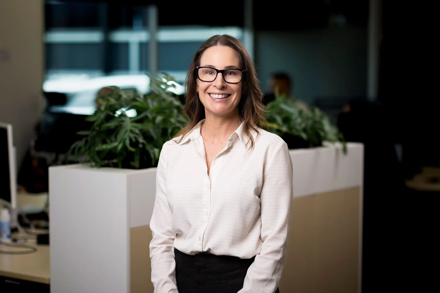 A smiling woman with glasses in an office setting, standing near plants and office furniture.