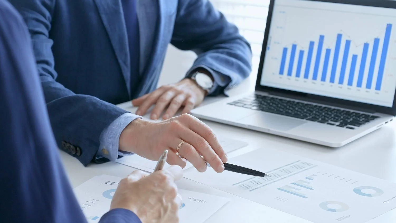 Two people in business suits analyzing financial documents with a laptop displaying a bar chart showing upward trends