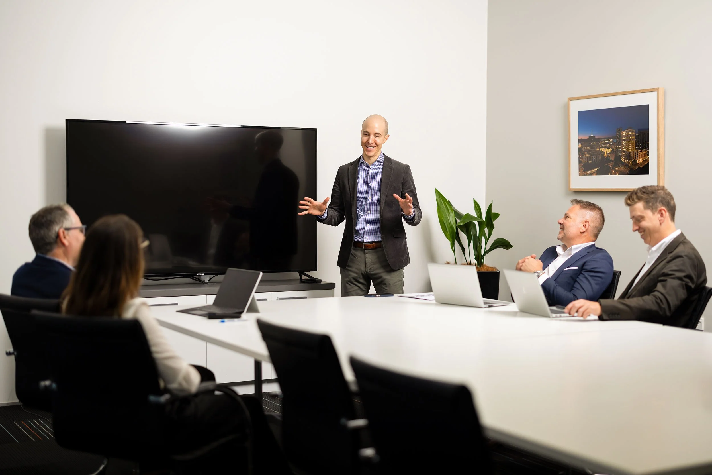 A man in a blazer and shirt giving a presentation to a group of people in a modern conference room with a large flat screen television, a framed cityscape photograph on the wall, and a potted plant.