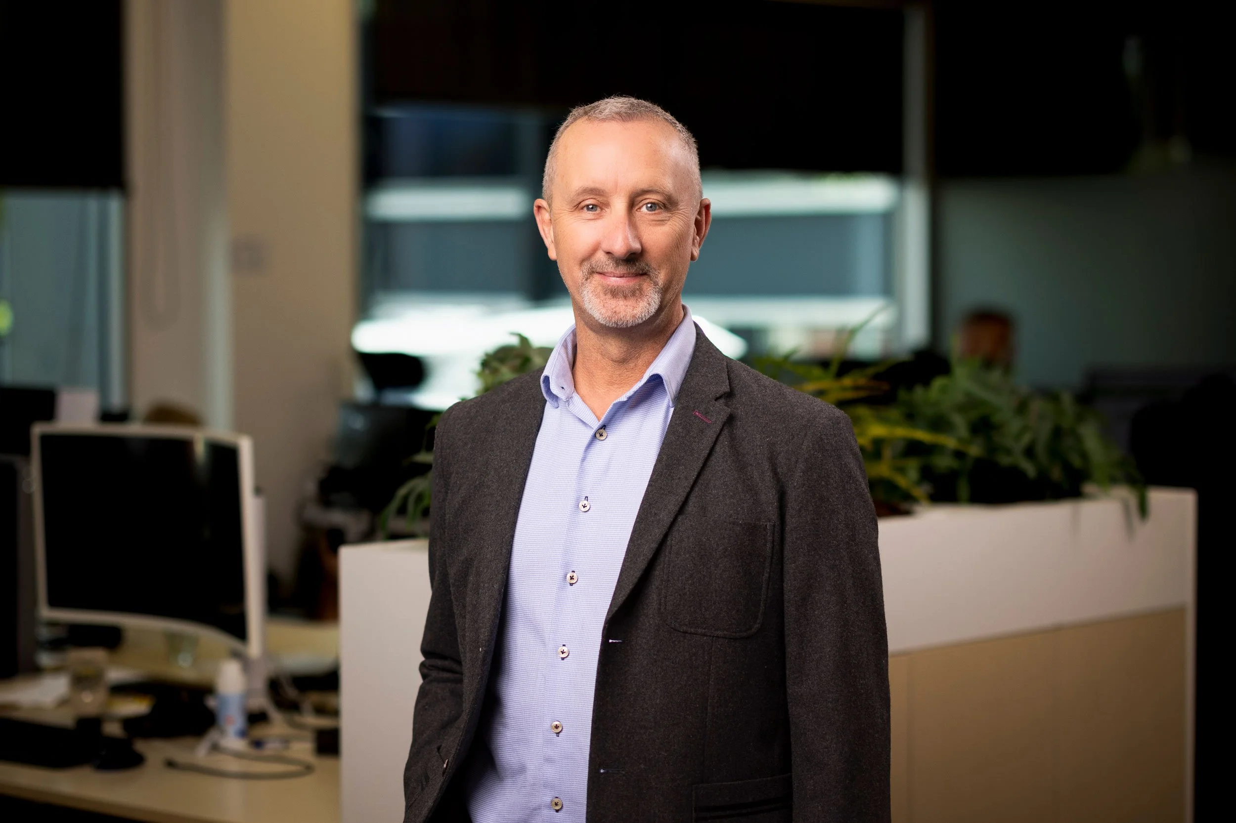 A middle-aged man with short gray hair and a beard, wearing a light blue dress shirt and dark blazer, standing in an office environment with computer desks and plants in the background.