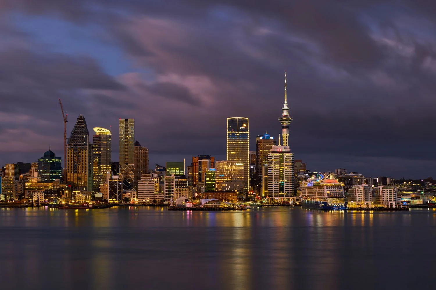 Nighttime view of a city skyline with illuminated buildings and a tall tower, reflecting on the water in the foreground, under a cloudy sky.