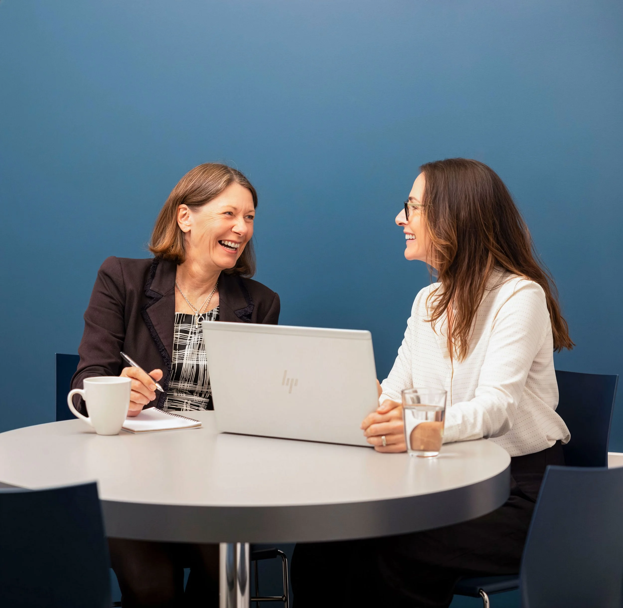 Two women are smiling and engaging in conversation at a round white table in an office room with a blue wall. One woman has shoulder-length gray hair, wears a black blazer and patterned blouse, holds a pen and notes, and has a coffee mug in front of her. The other woman has long brown hair, wears glasses and a white blazer, and has a glass of water in front of her.