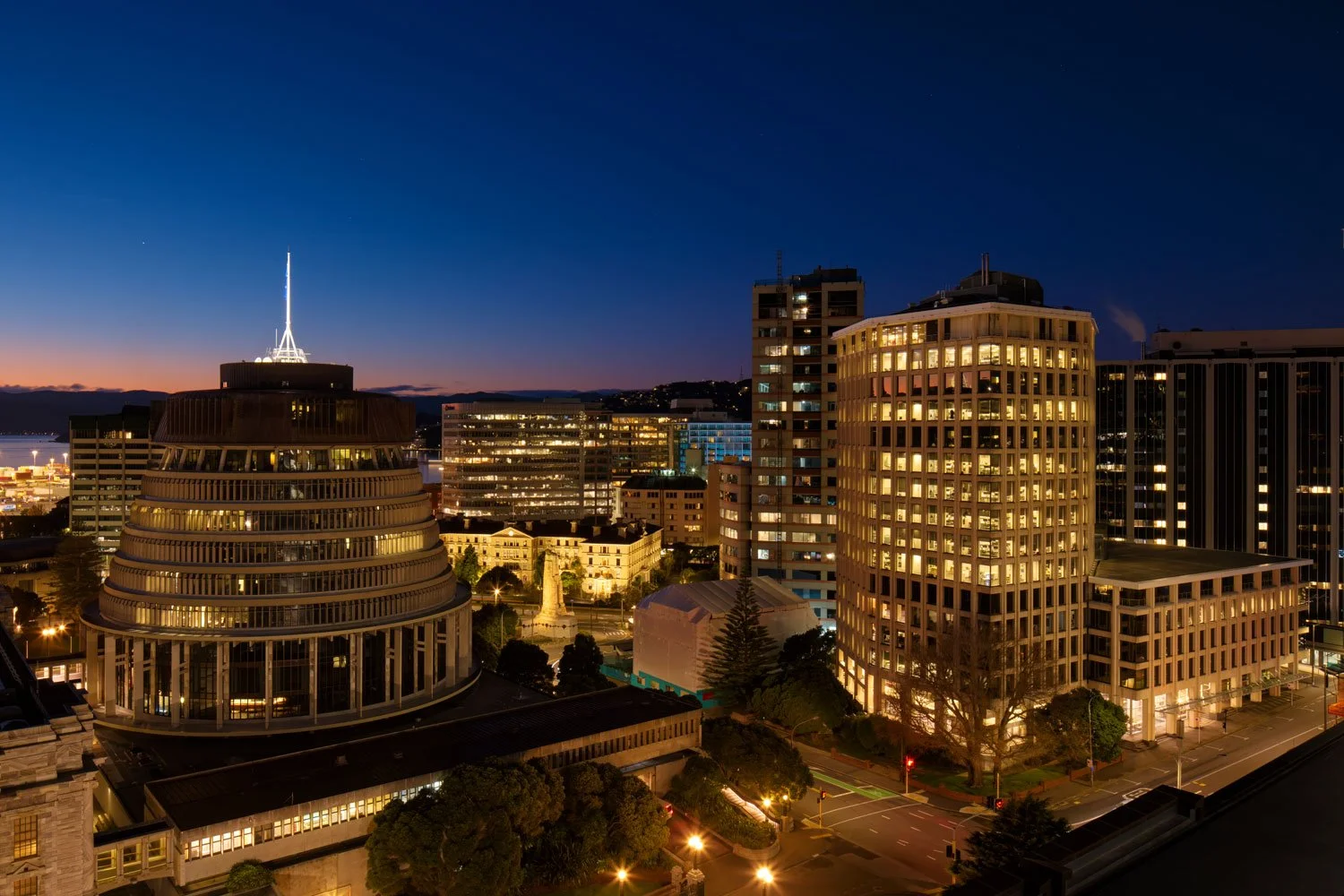 Nighttime cityscape featuring illuminated buildings, with a clear sky and a gradient from orange to deep blue, and water in the distance.