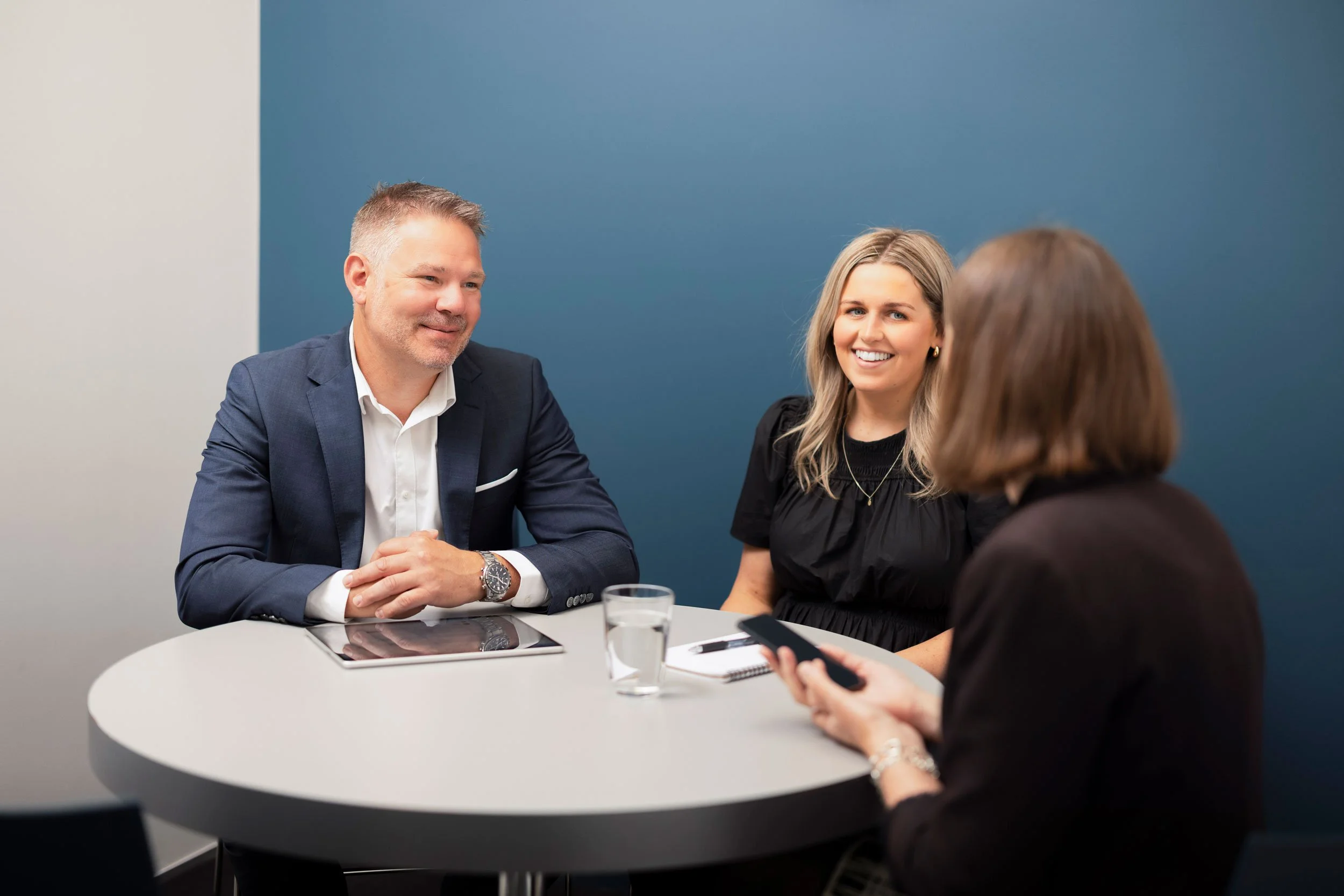A group of four people sitting around a white round table in a meeting room with a blue wall, engaged in conversation. One man in a dark suit and white shirt is smiling, while a woman with blonde hair and a black top in front of him is also smiling. Two other women, one with brown hair, are partially visible, one holding a smartphone. Items on the table include a tablet, pen, notebook, and a glass of water.