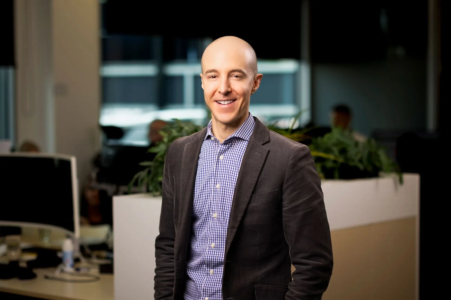 A smiling bald man in a dark blazer and checkered shirt standing in an office environment.