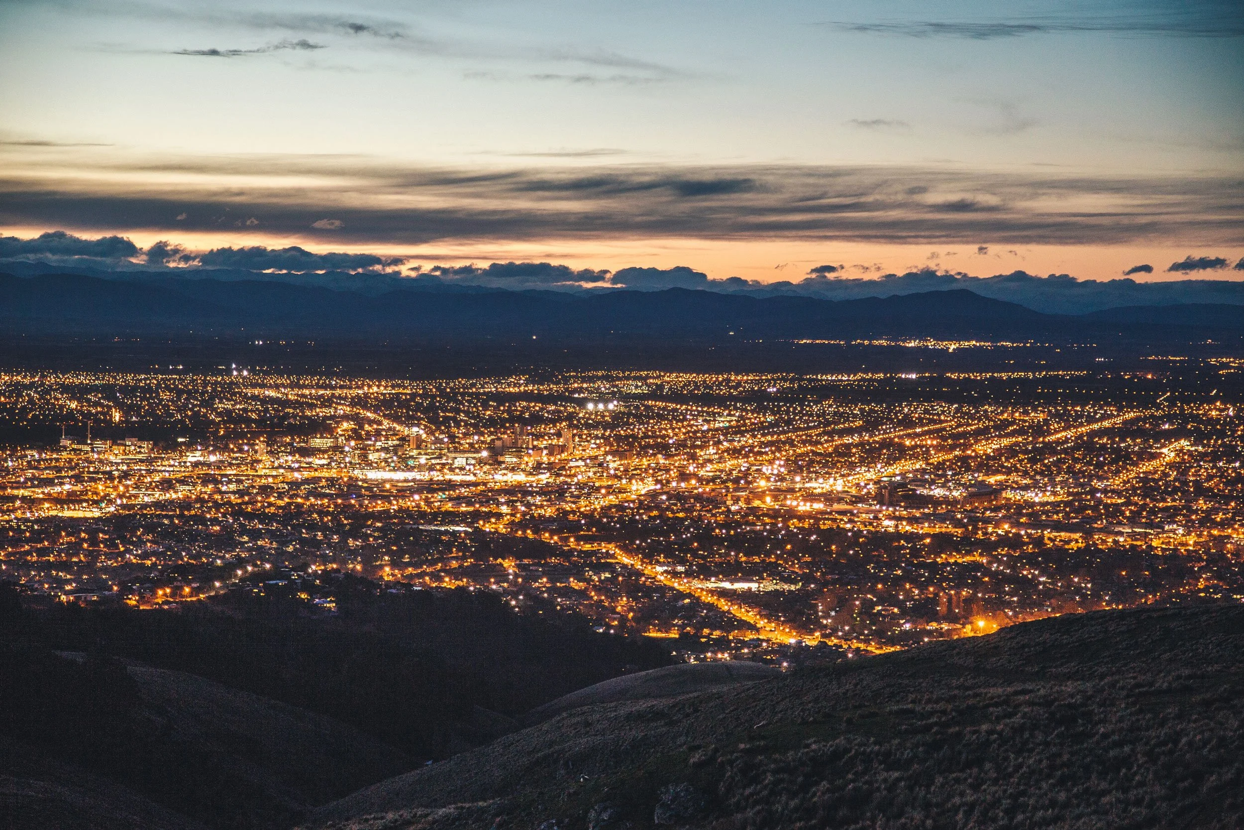 Nighttime cityscape viewed from a high vantage point with illuminated streets and a mountain range in the background.